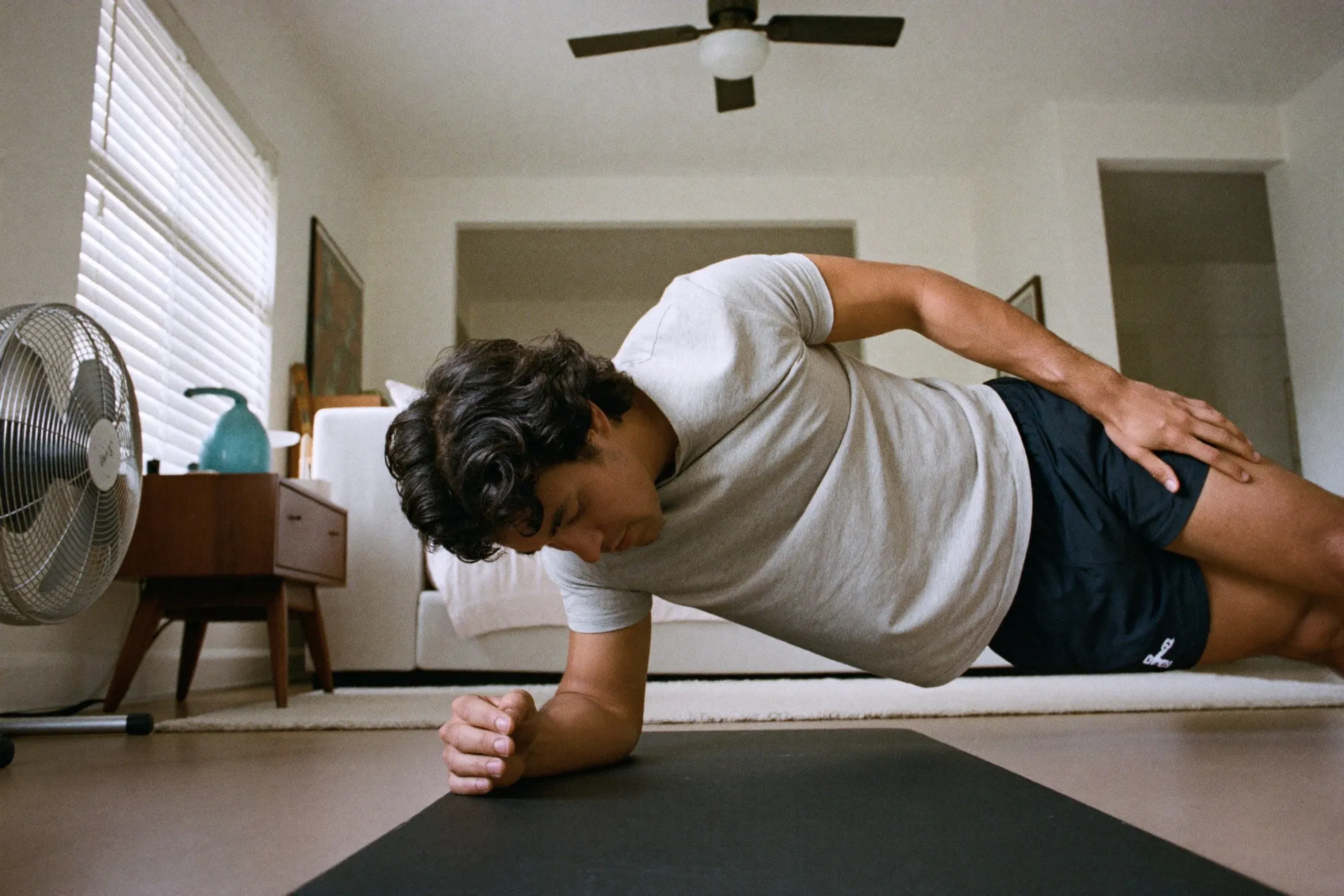 Man holding a forearm plank on an exercise mat in a bright HDB flat living room, morning light through the window
