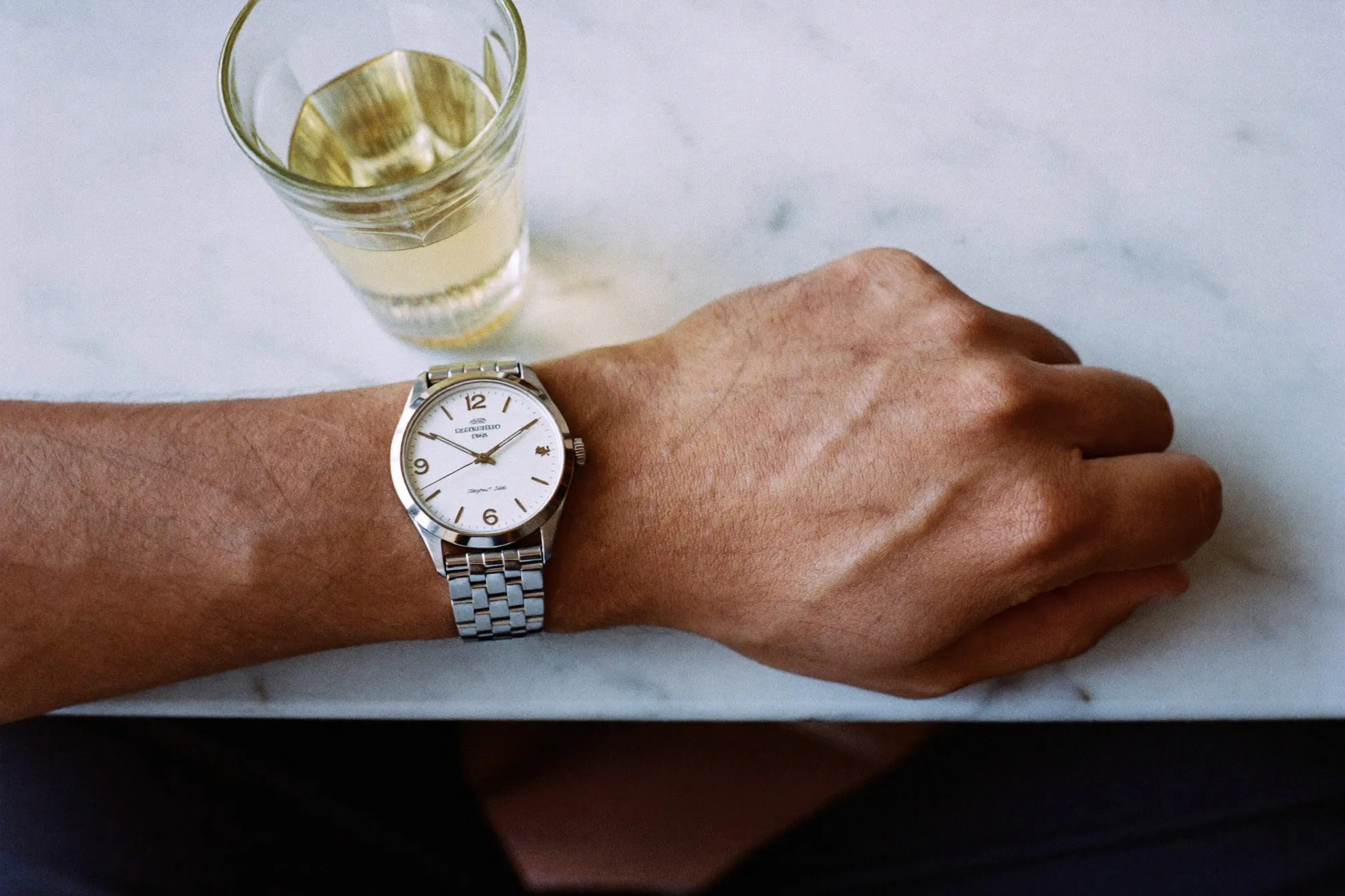 Close view of a man's wrist with a plain steel watch resting on a kopitiam marble table beside a glass of iced coffee