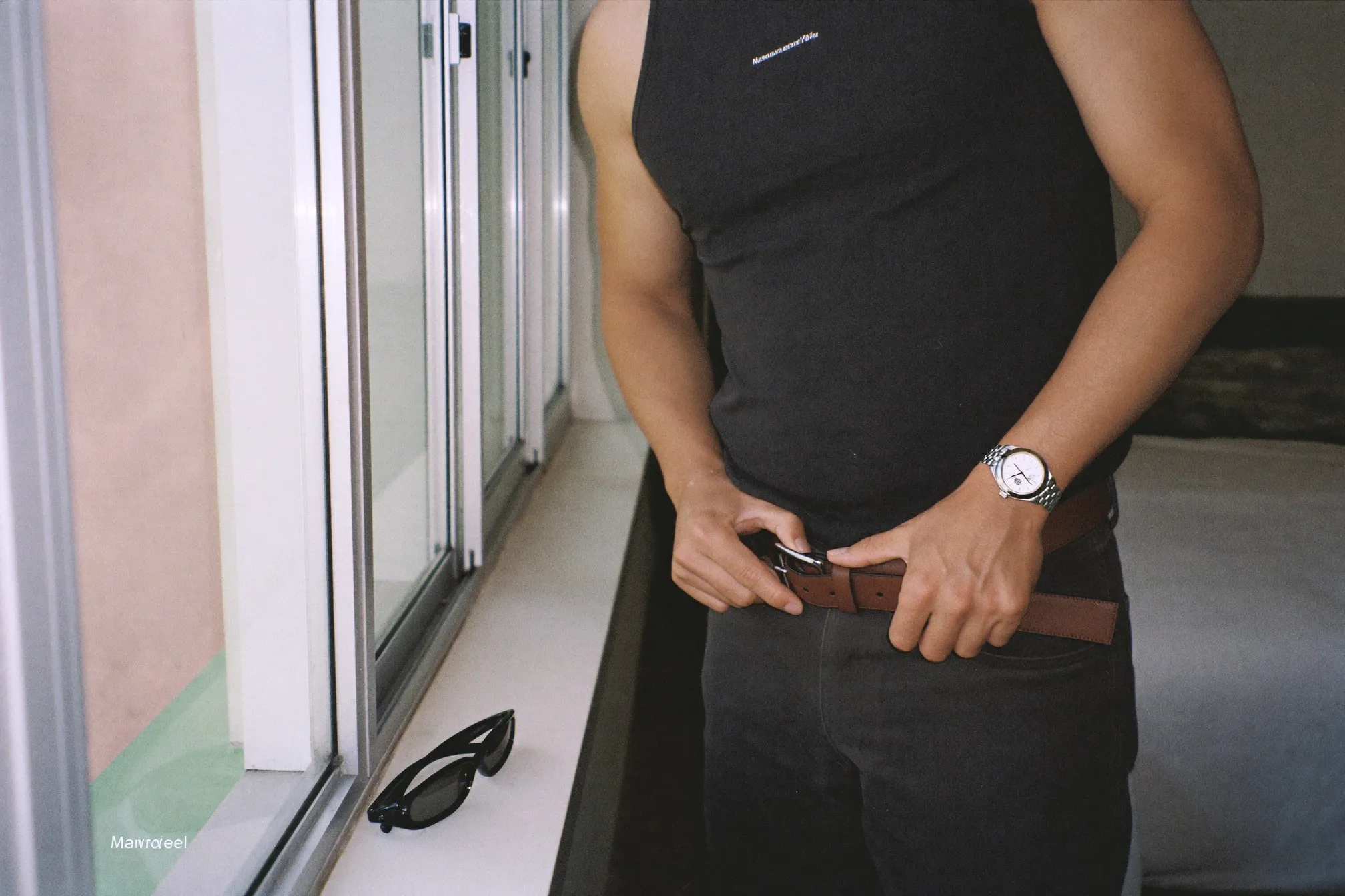 Man fastening a brown leather belt at a bedroom window, plain steel watch on his wrist and sunglasses resting on the sill
