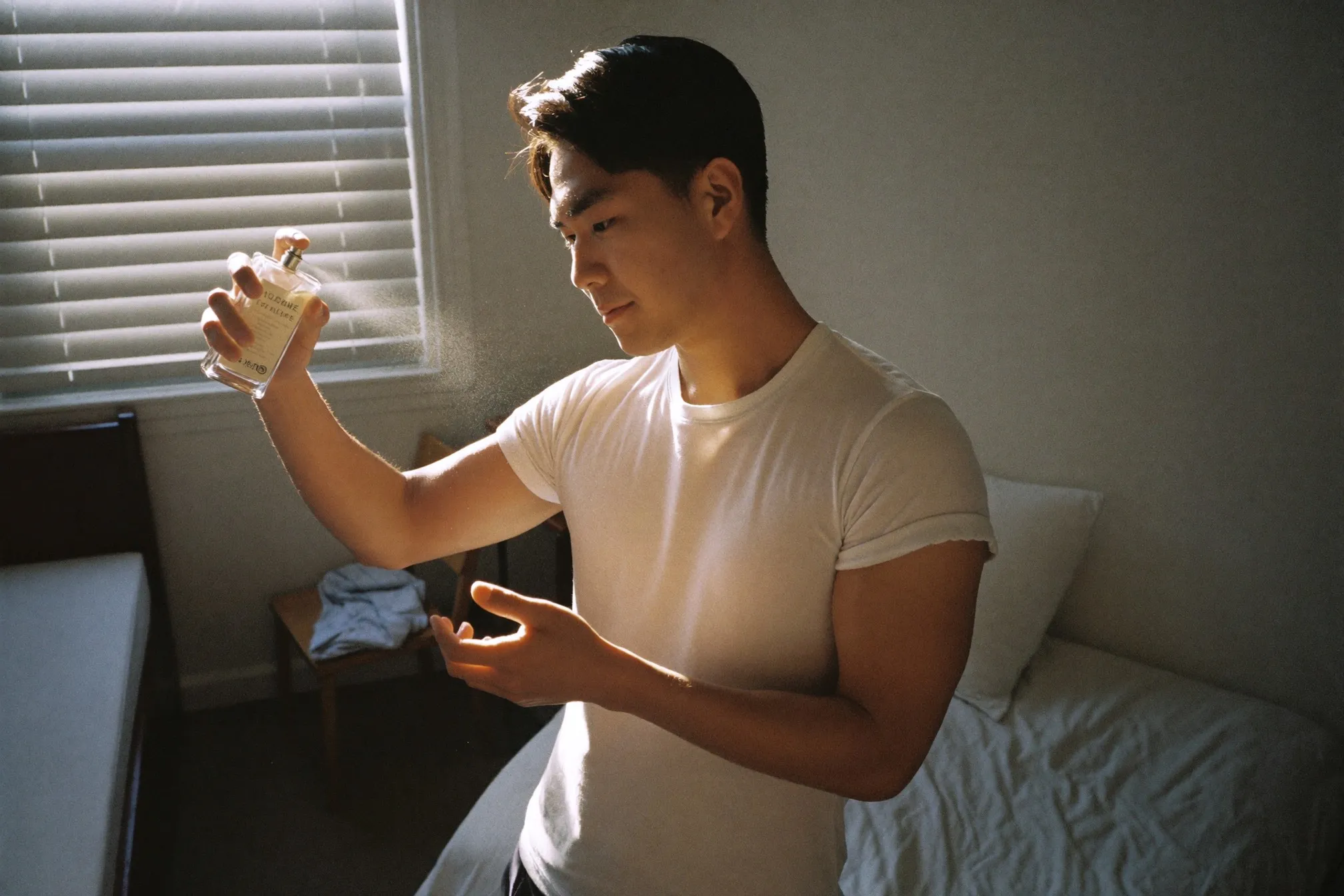 Man in a white t-shirt spraying an unbranded glass fragrance bottle onto his forearm in an HDB bedroom in the morning