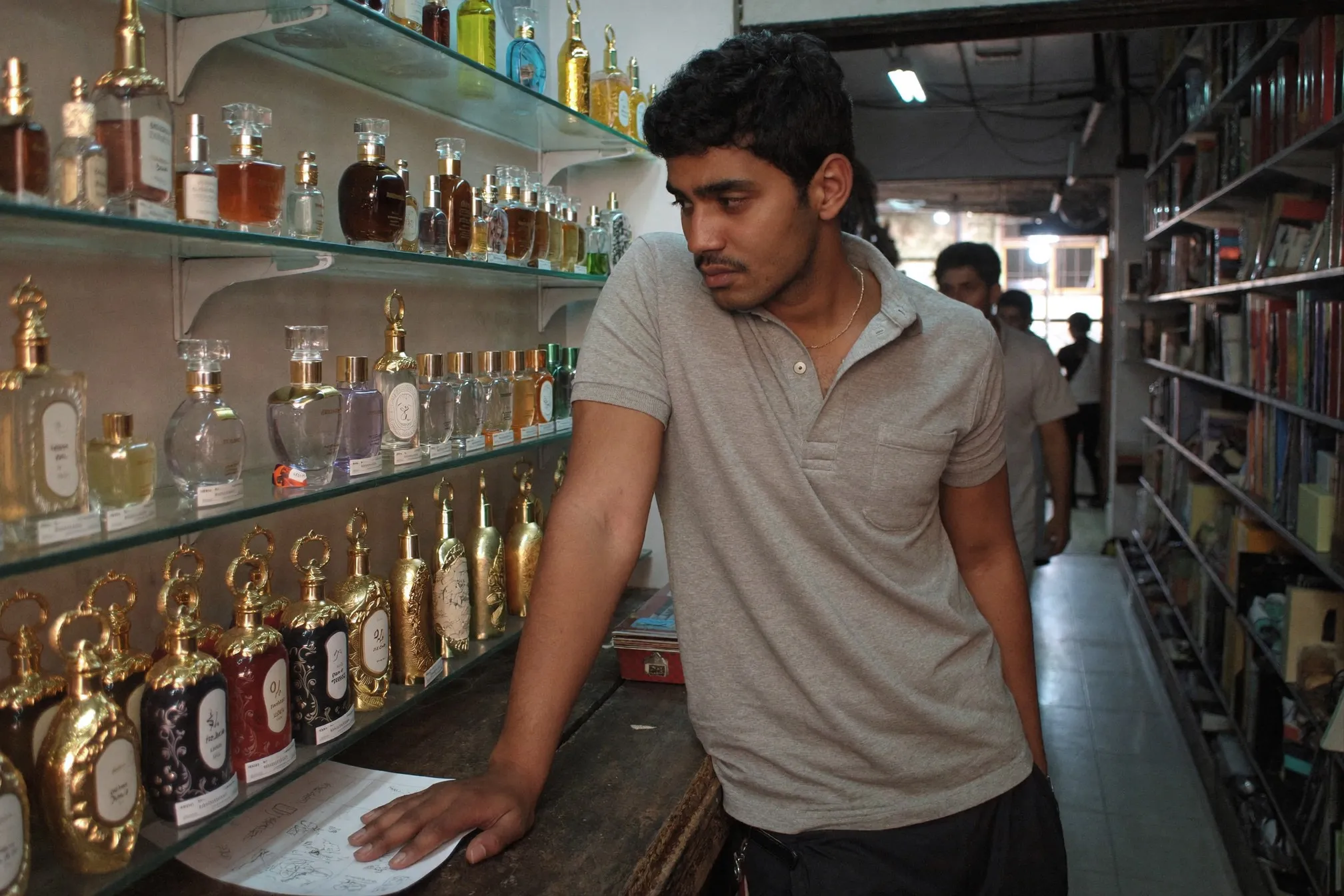 Man at a small perfume shop counter in Kuala Lumpur looking at rows of unbranded ornate glass bottles on a lit glass shelf