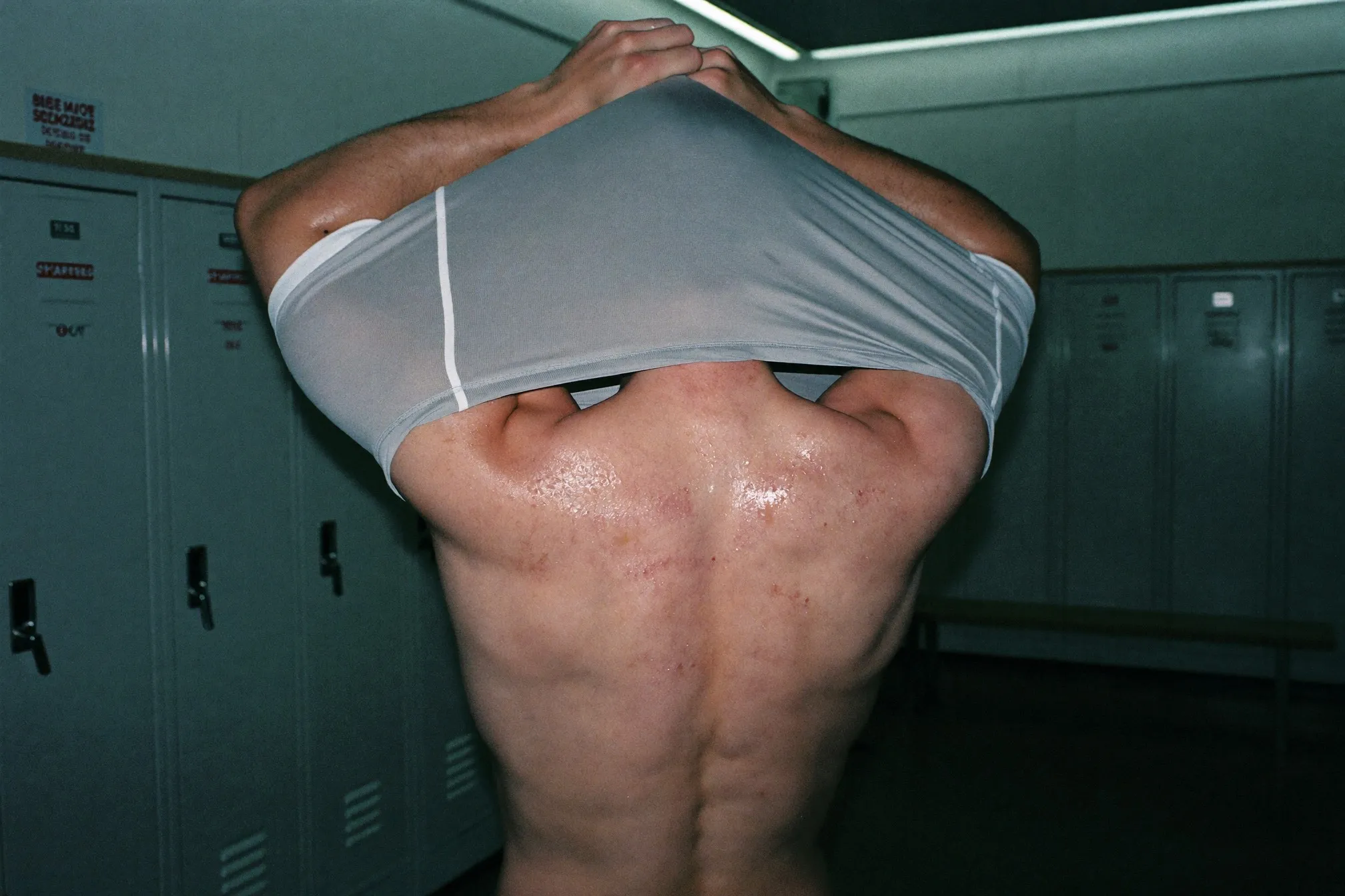 Man pulling a sweat-soaked training shirt over his head in a gym changing room