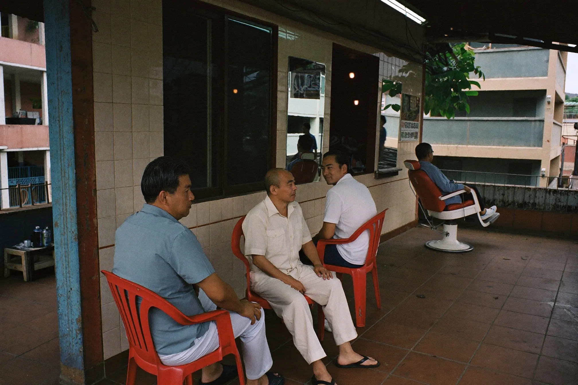 Men waiting their turn on plastic chairs at a neighbourhood barbershop under an HDB block