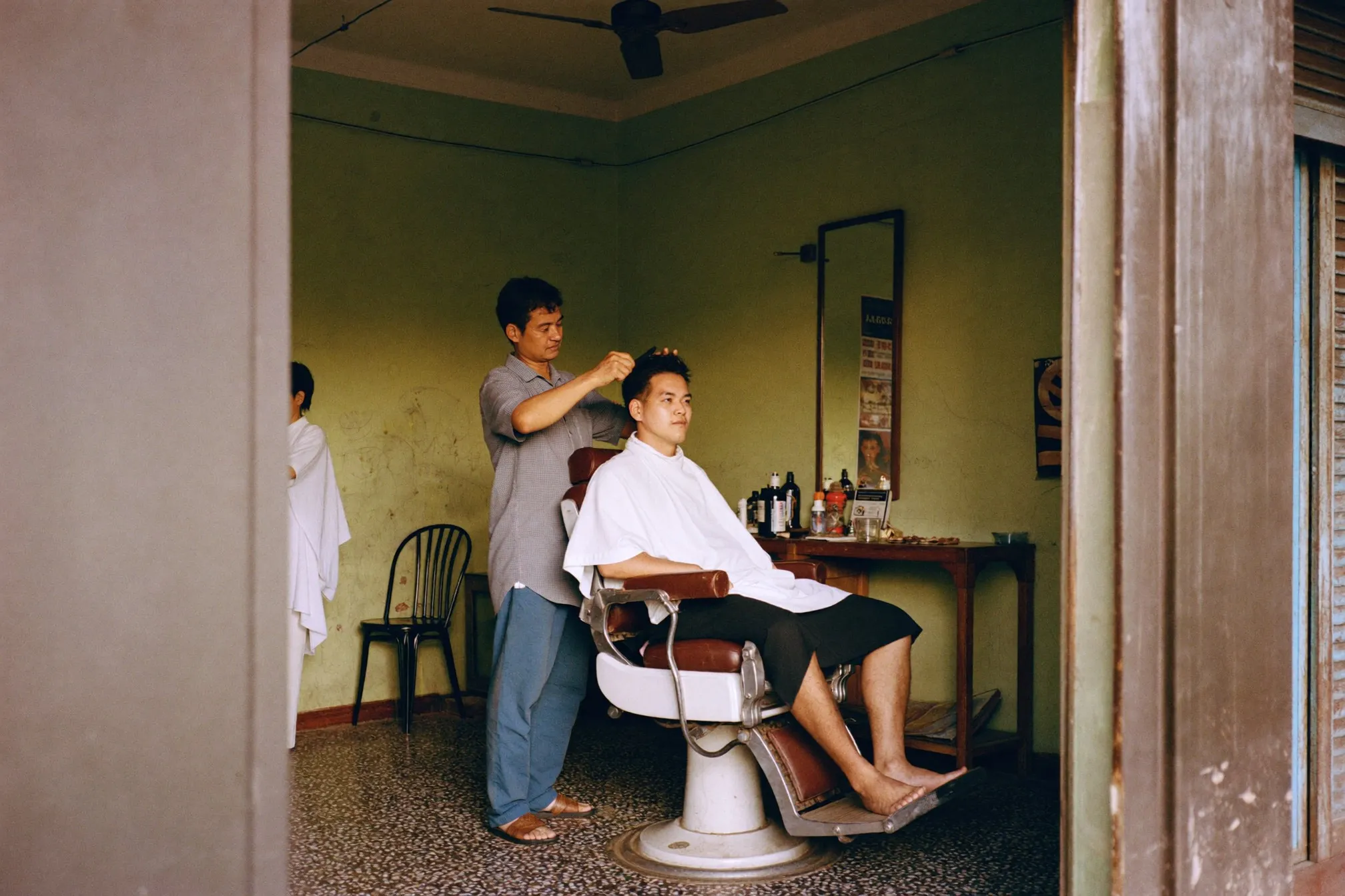 Barber cutting a seated customer's hair in an old shoplot barbershop in Malaysia with a ceiling fan overhead