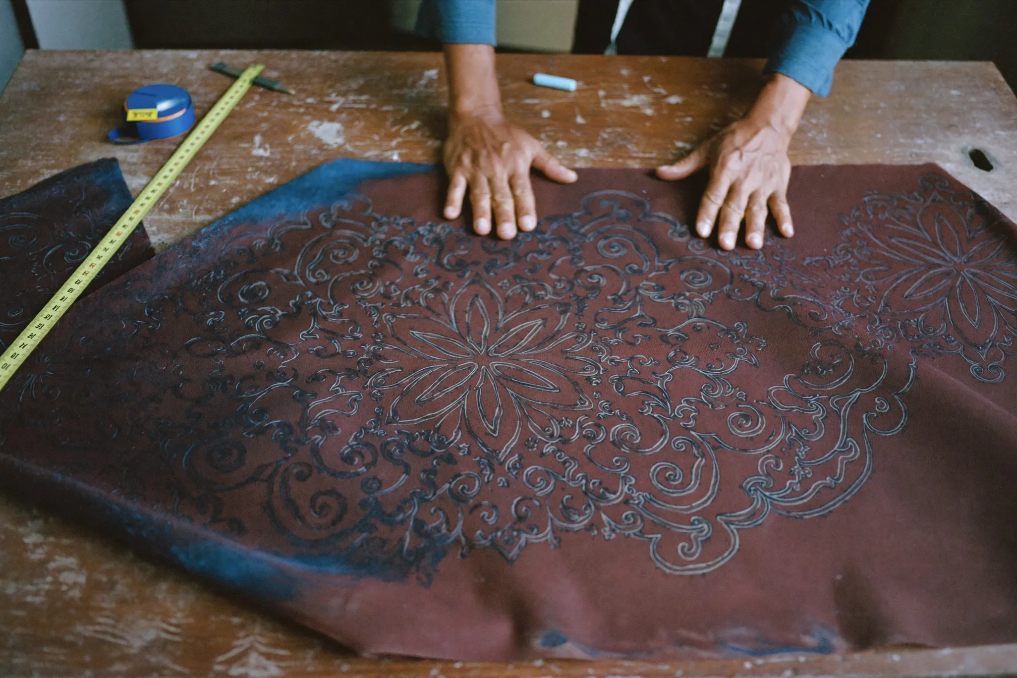 Tailor's hands smoothing hand-painted batik cloth with large floral motifs across a wooden cutting table