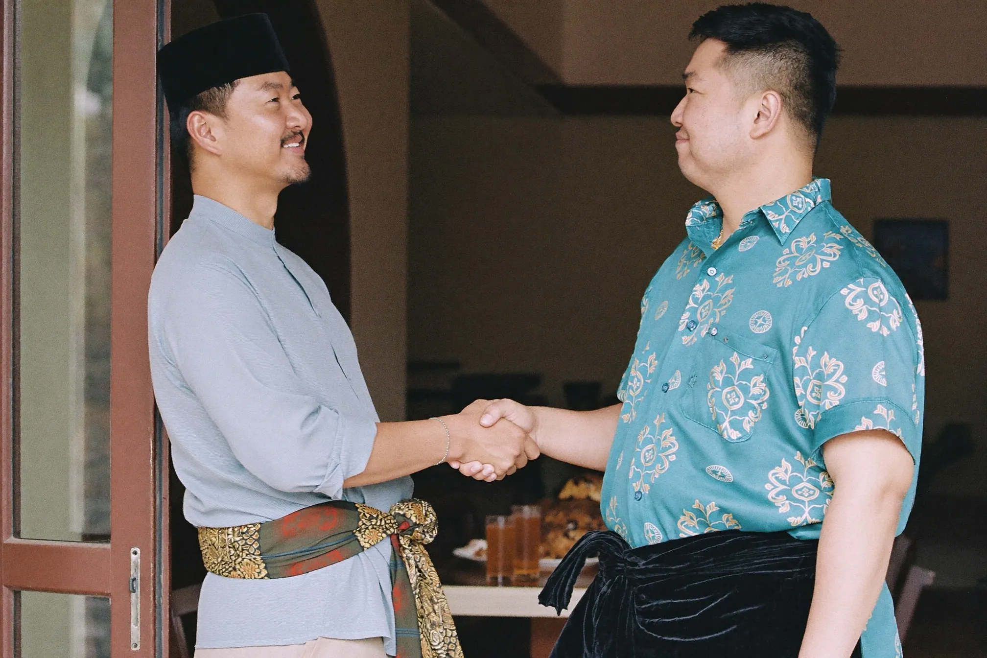 Man in a light blue baju melayu with songket sampin and black songkok greeting a guest in a batik shirt at a Hari Raya open house