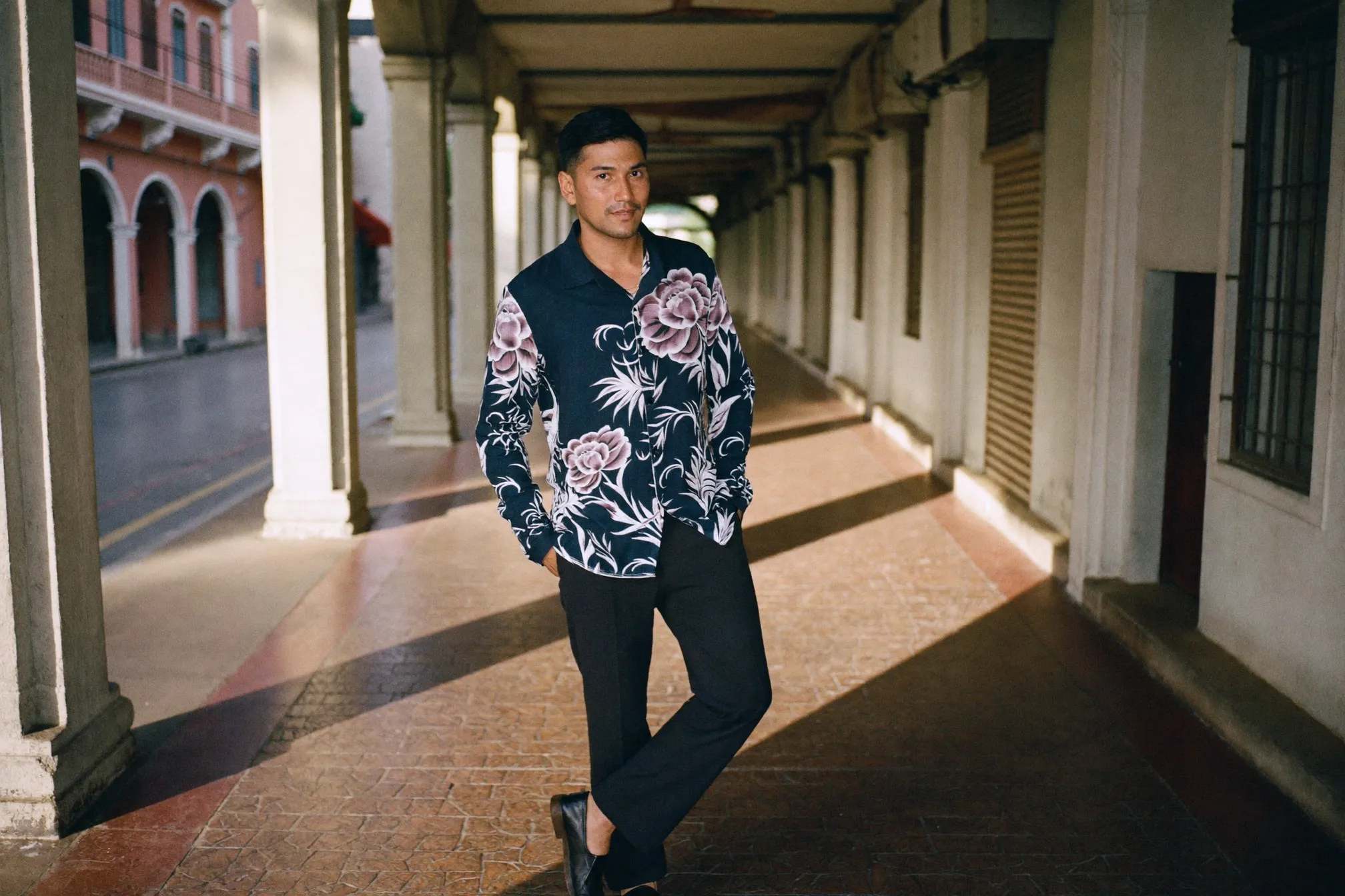 Malay man in a long-sleeved dark batik shirt and charcoal trousers standing in a shaded colonnade in Kuala Lumpur