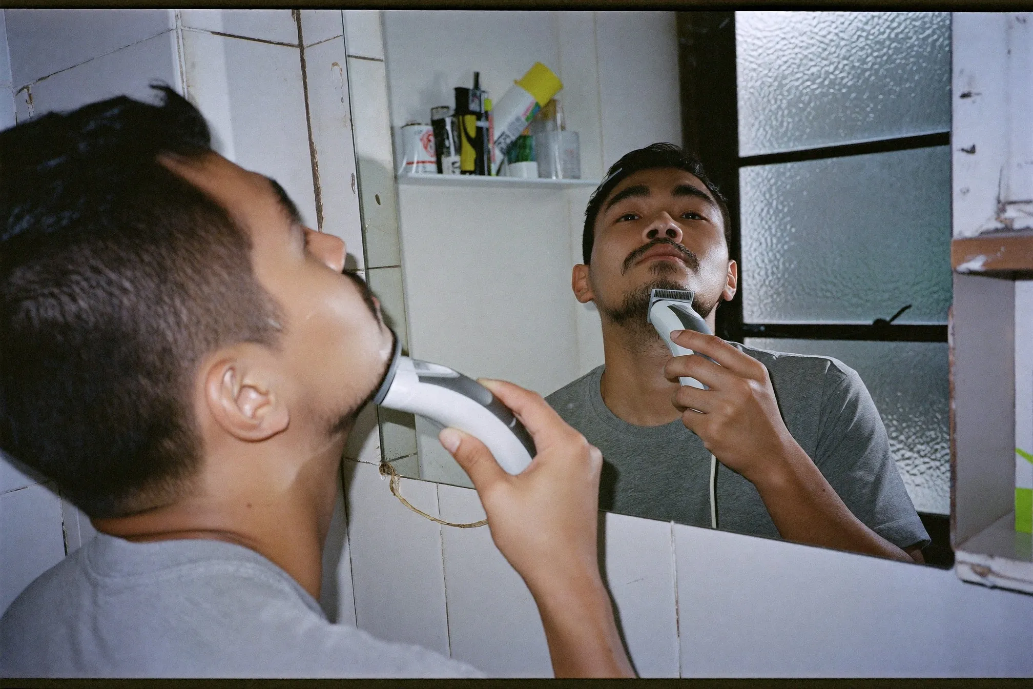 Man trimming his short beard with a cordless trimmer at a bathroom mirror in the morning