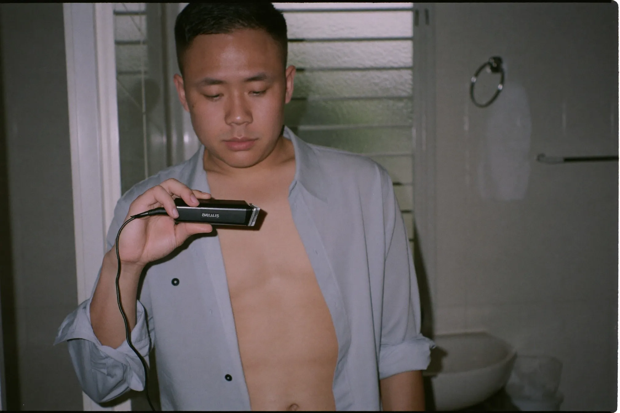 Man in an open plain cotton shirt standing at a bathroom mirror holding a cordless body trimmer