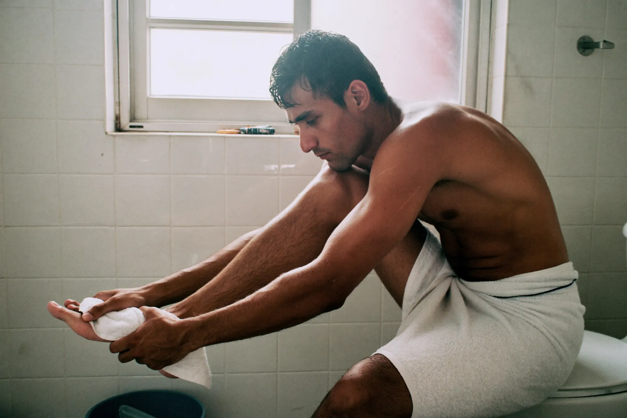 Man drying between his toes with a towel on the edge of a small bathroom