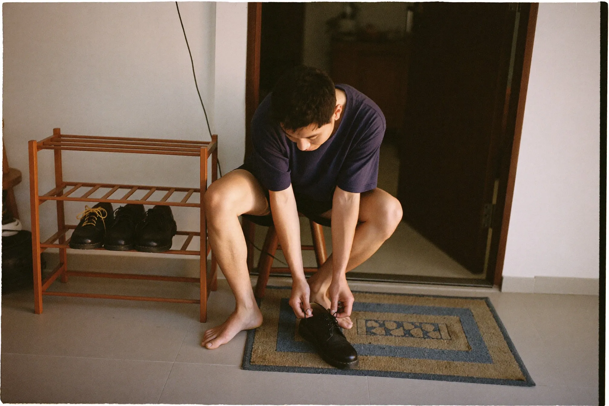 Man sitting on a low stool at his apartment doorway unlacing leather work shoes beside a wooden shoe rack