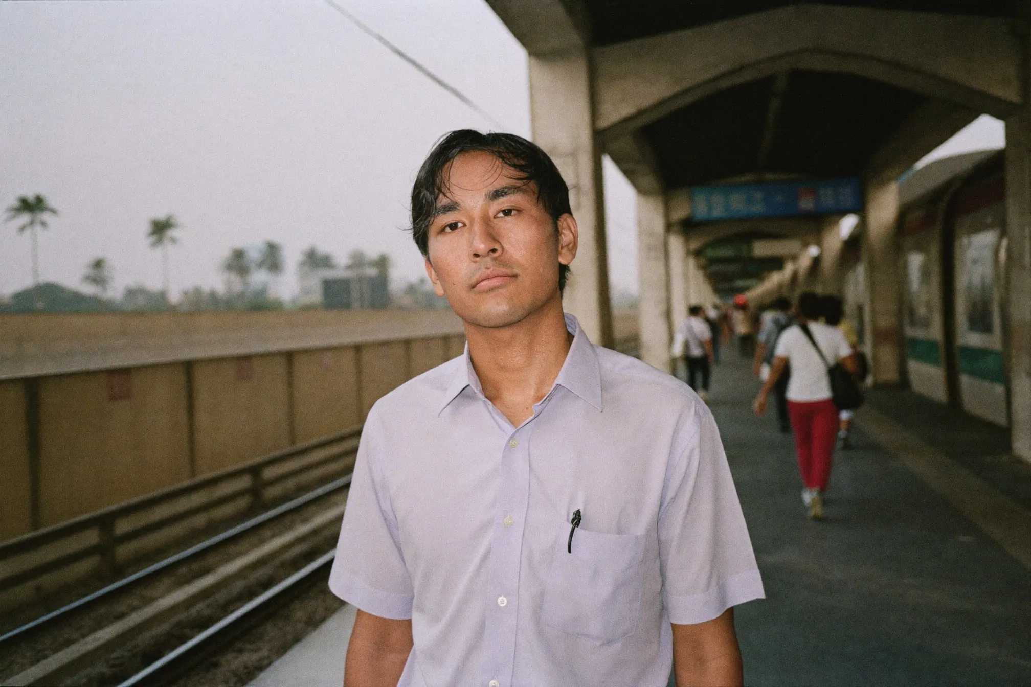 Man waiting on a crowded open-air train platform in the late afternoon heat, shirt showing a faint sweat patch