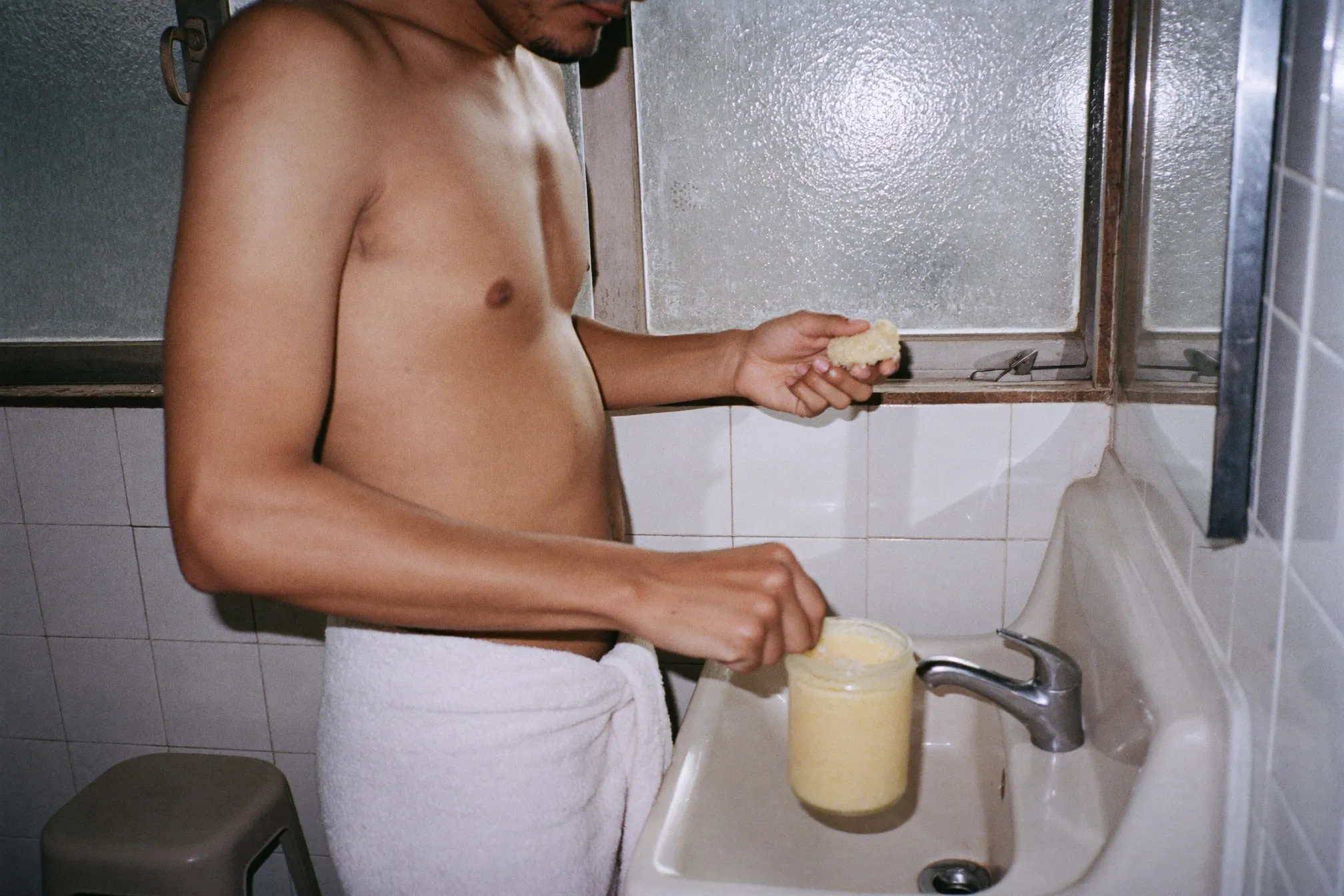 Man scooping a coarse scrub from an unlabelled glass jar beside a bathroom sink in a Singapore flat