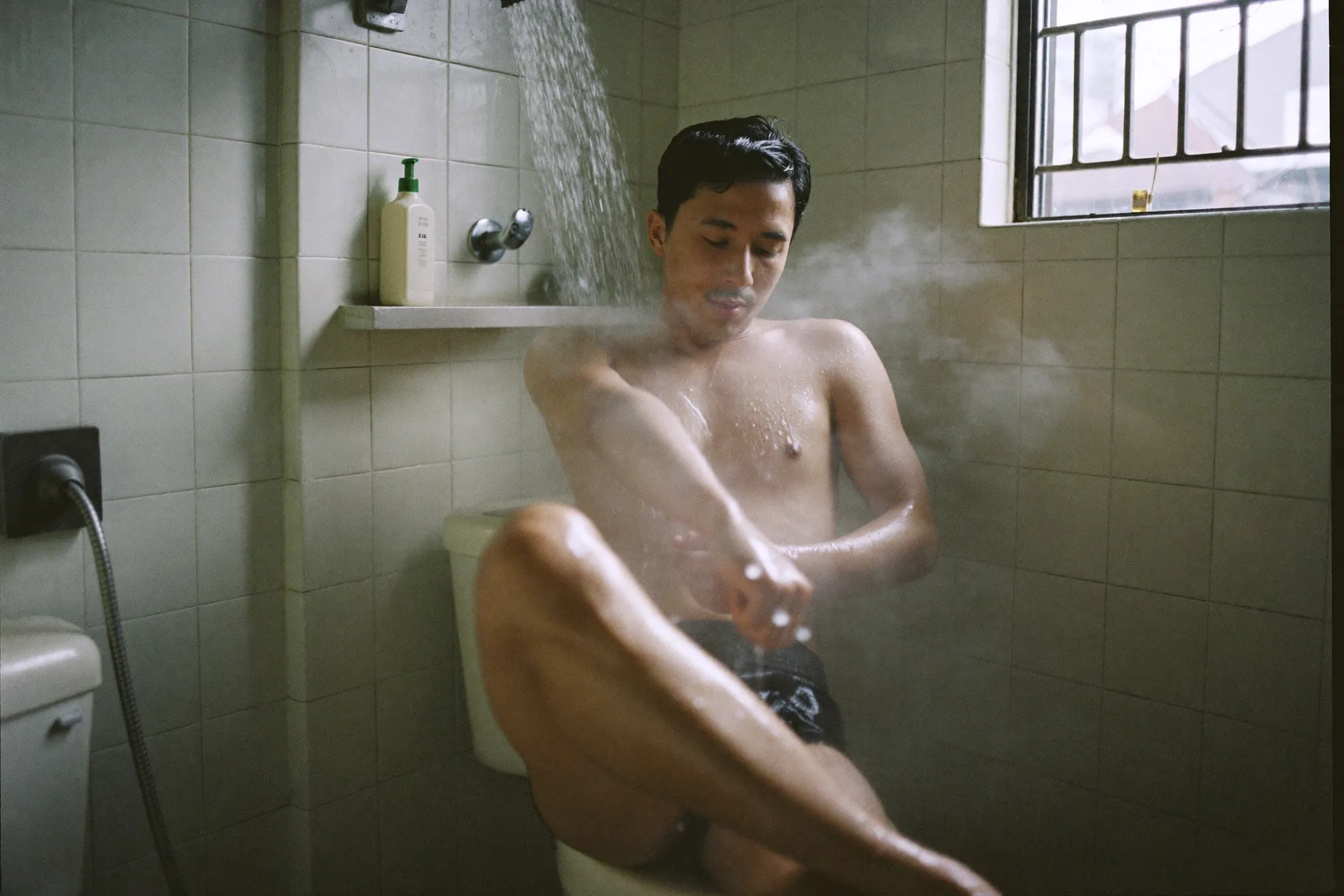 Man rinsing off in a small tiled bathroom in a Kuala Lumpur flat with morning light through a window grille