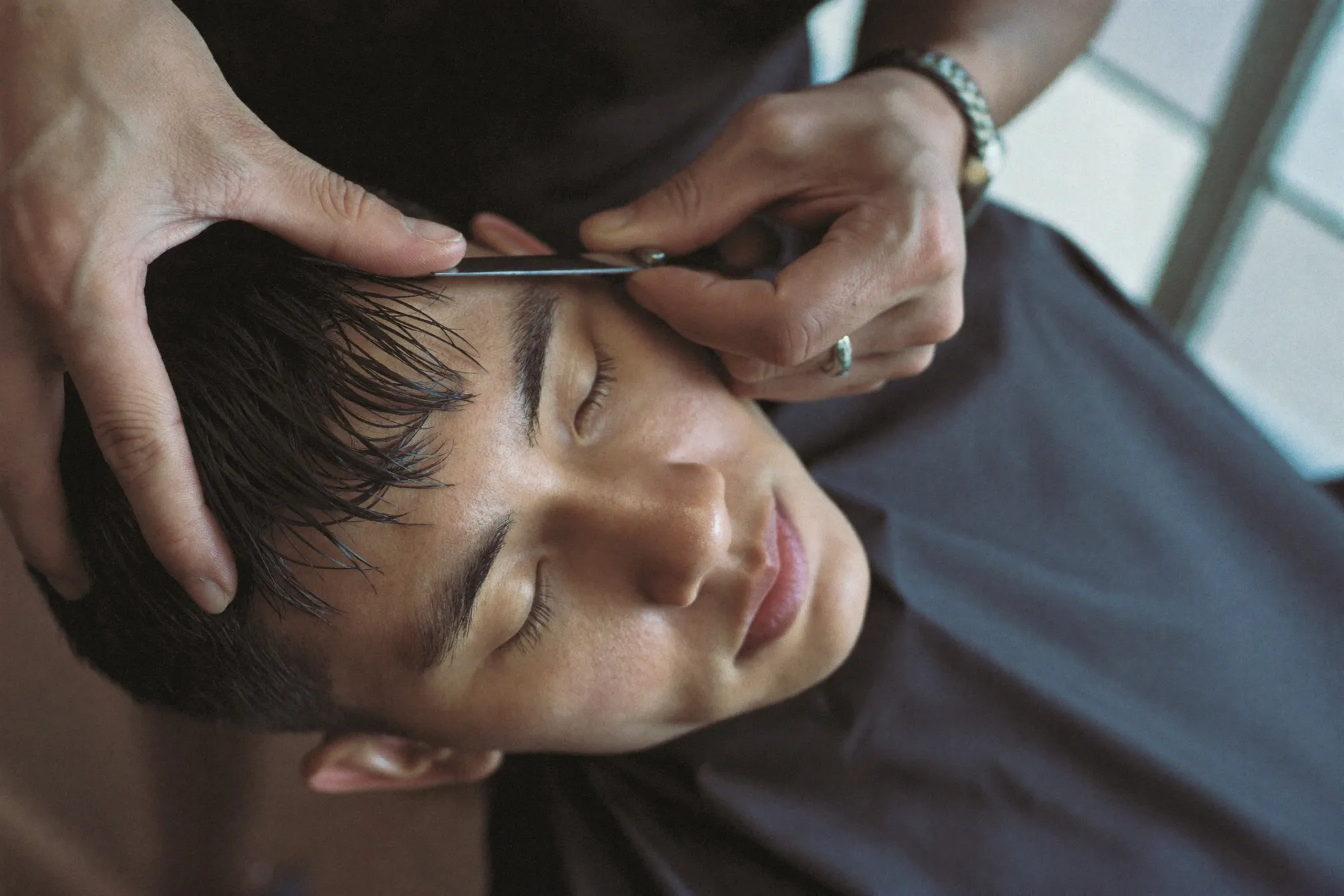 Barber point-cutting the fringe of a young Malay man's boyish haircut with scissors held vertically