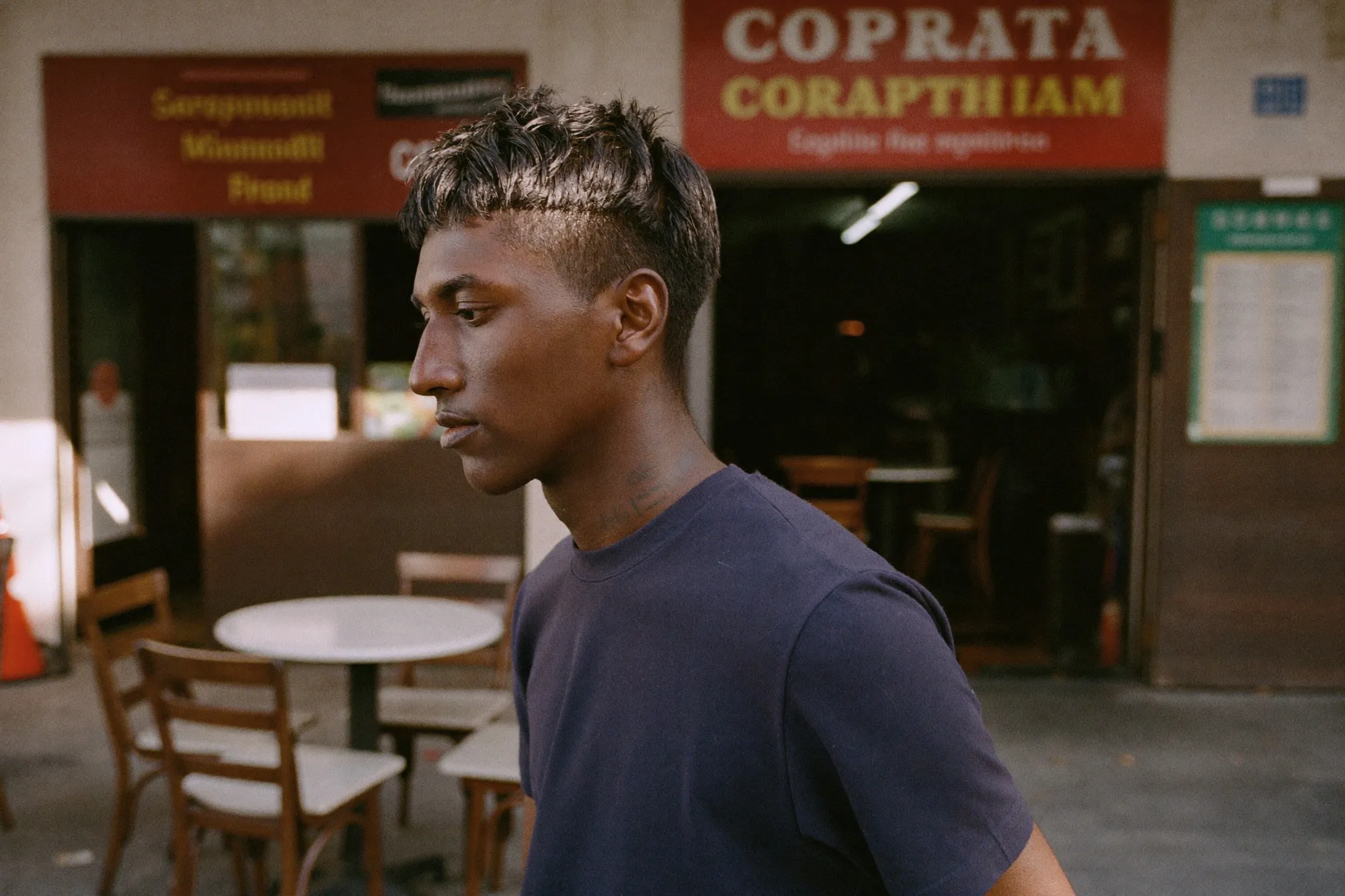Young Indian Singaporean man with a boyish haircut walking past a kopitiam in the late afternoon
