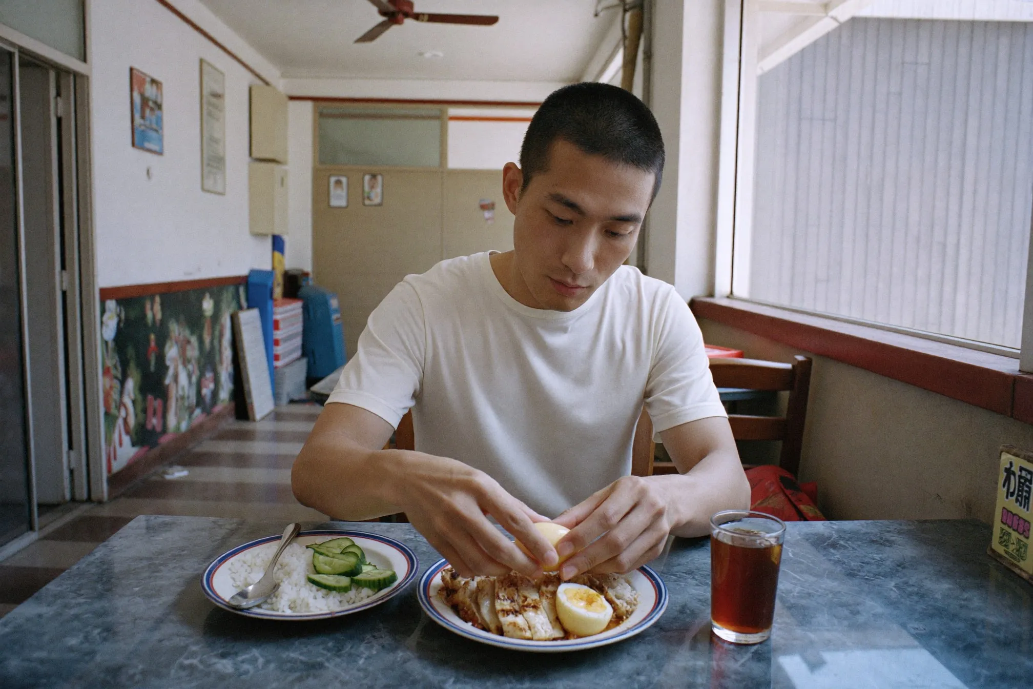 Chinese Malaysian man at a kopitiam table adding a hard-boiled egg to a plate of rice and grilled chicken