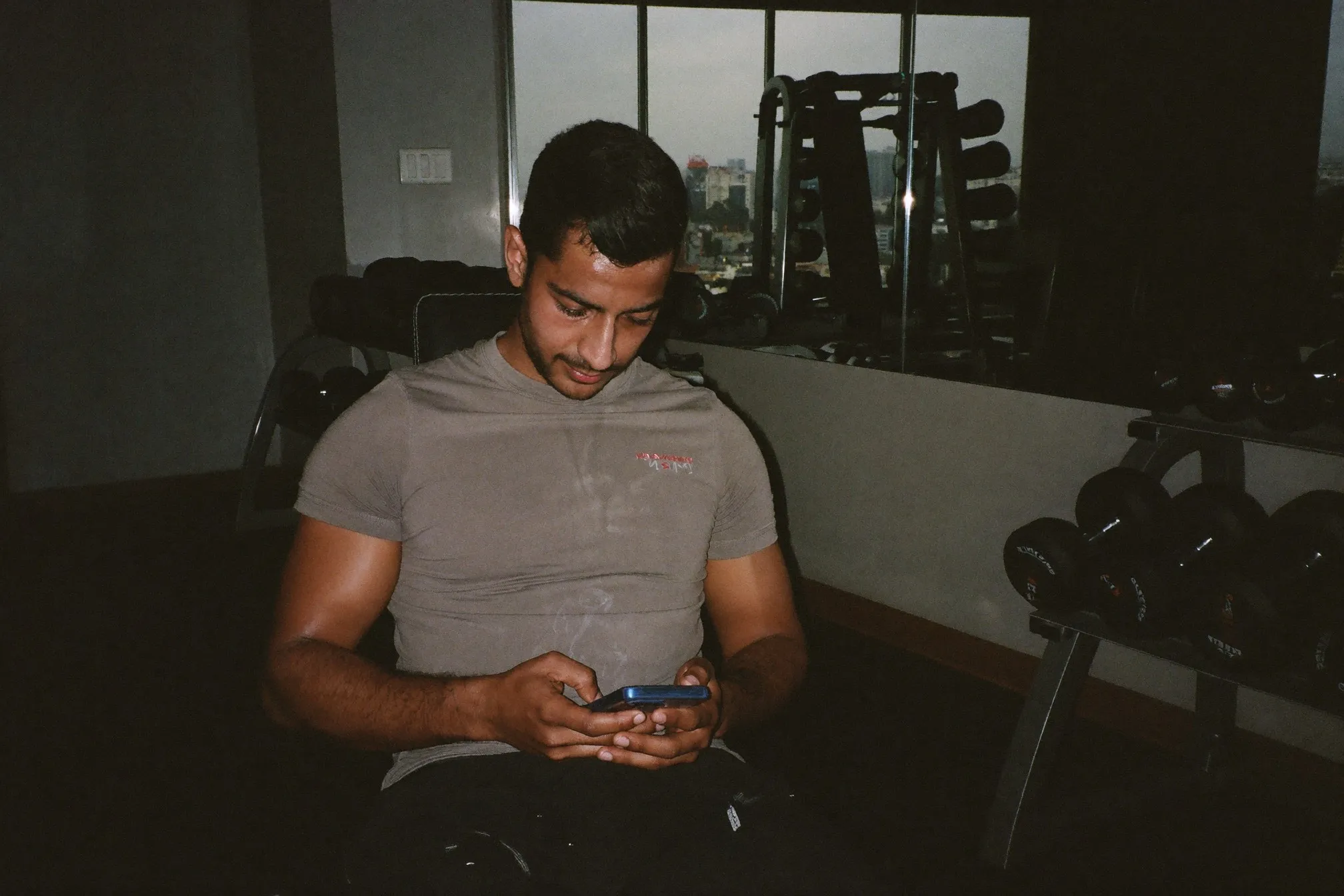 Indian man writing his sets into a phone notes app between dumbbell rows in a small condo gym