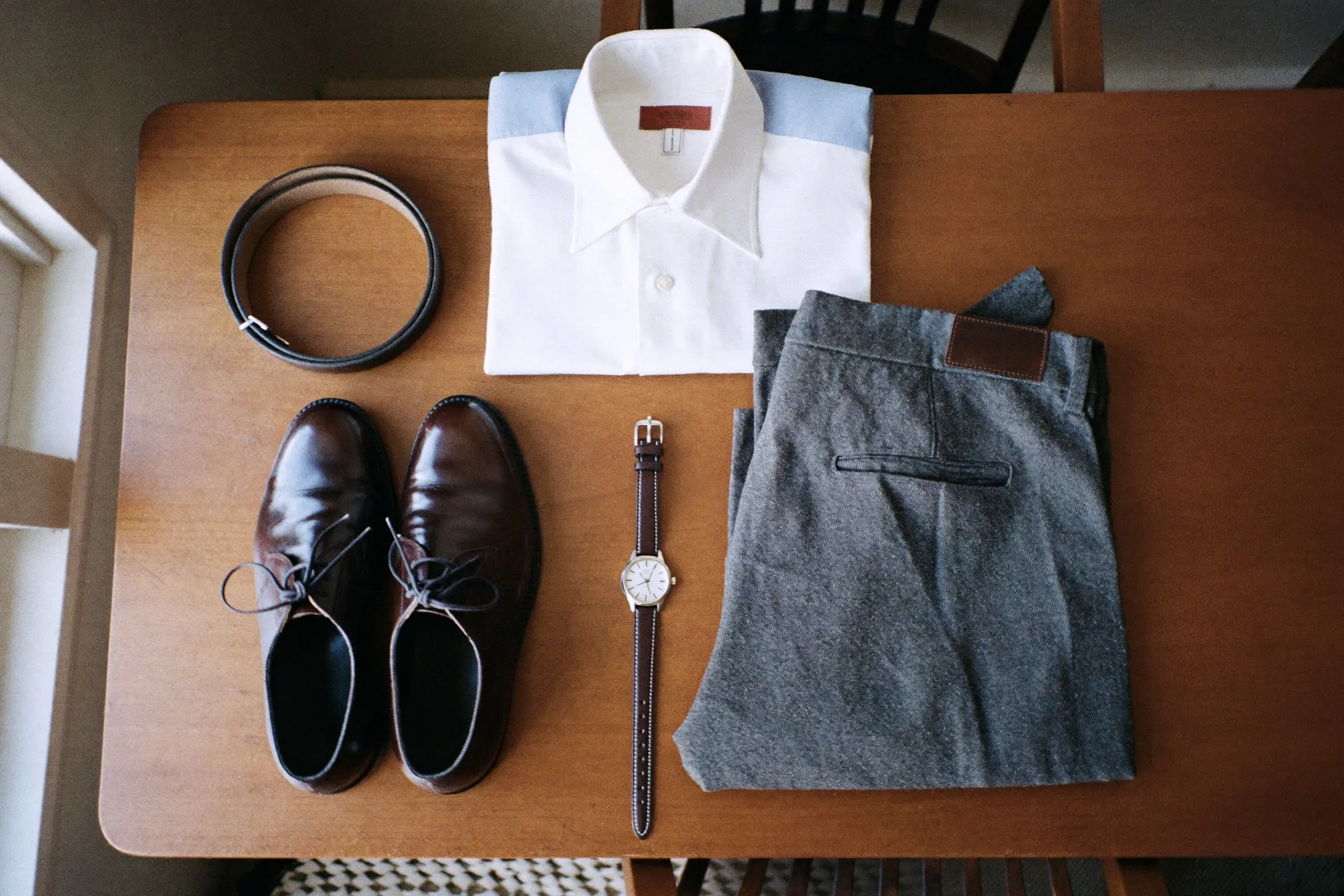 Flat lay of a business casual capsule: folded shirts, pressed grey trousers, brown derby shoes, a leather belt and a plain steel watch on a wooden desk