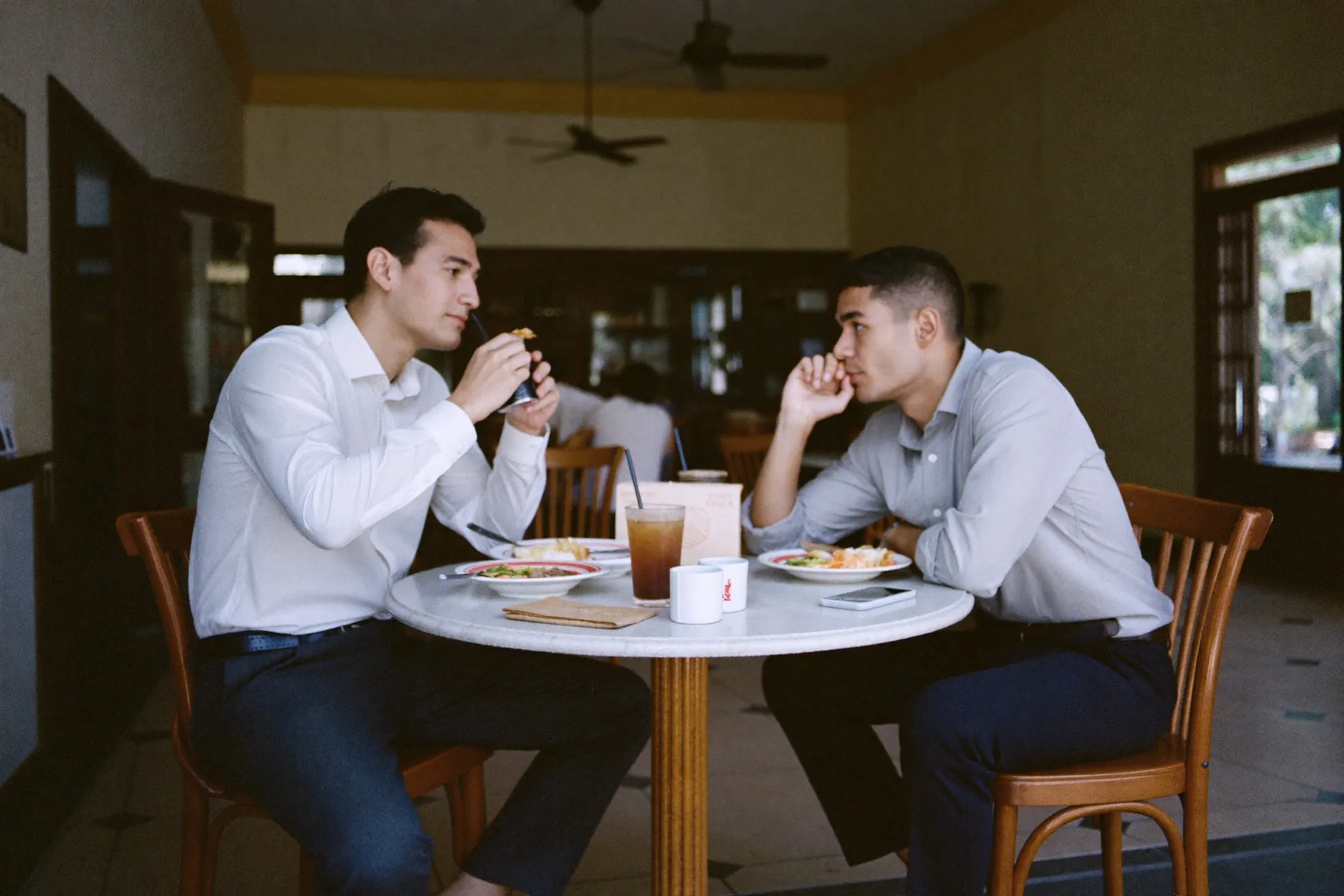 Two colleagues in long-sleeve shirts and dark trousers having lunch at a kopitiam marble table in Kuala Lumpur