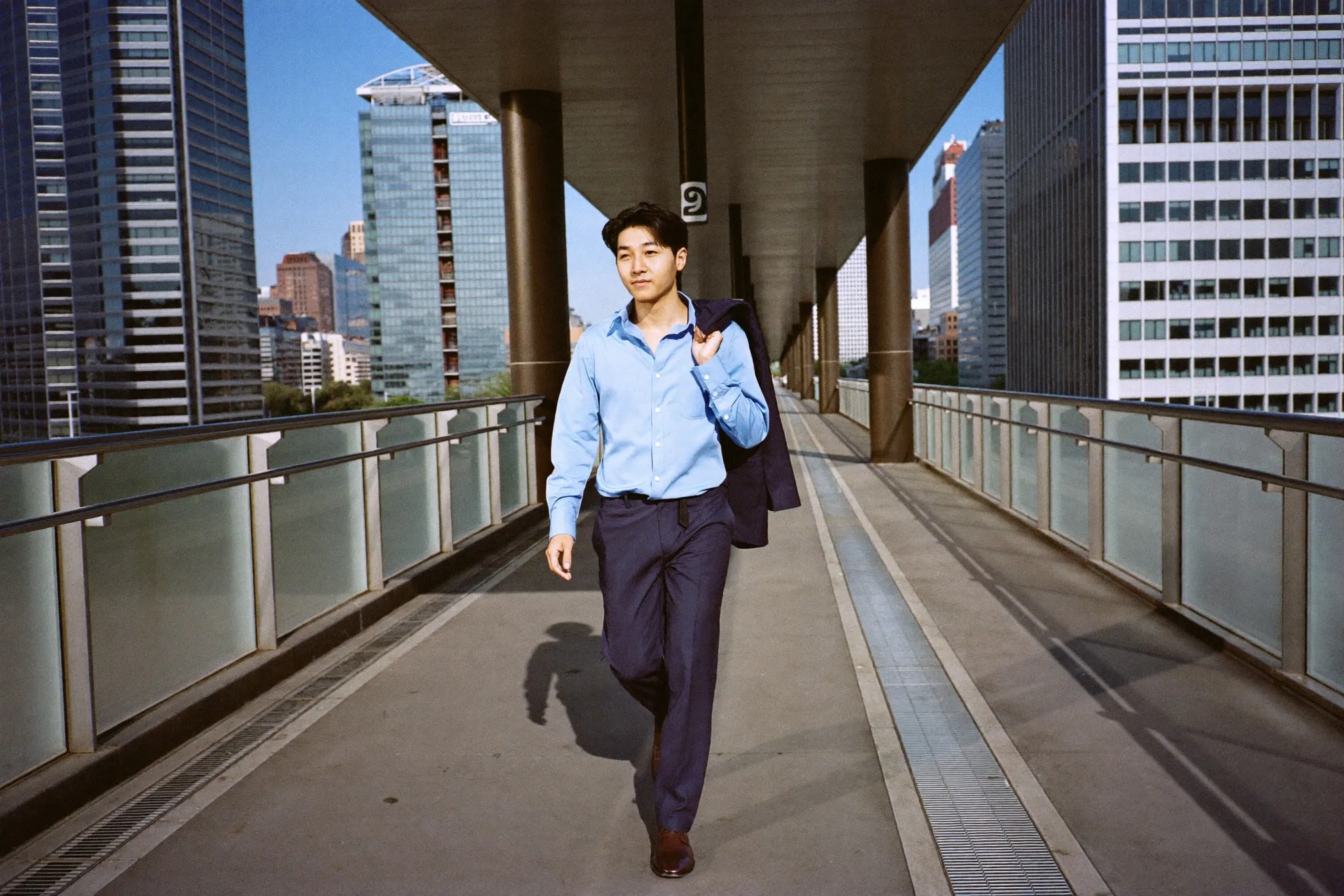 Man in a light blue long-sleeve shirt and navy trousers carrying a blazer over his shoulder on a covered walkway between office towers in Singapore's CBD