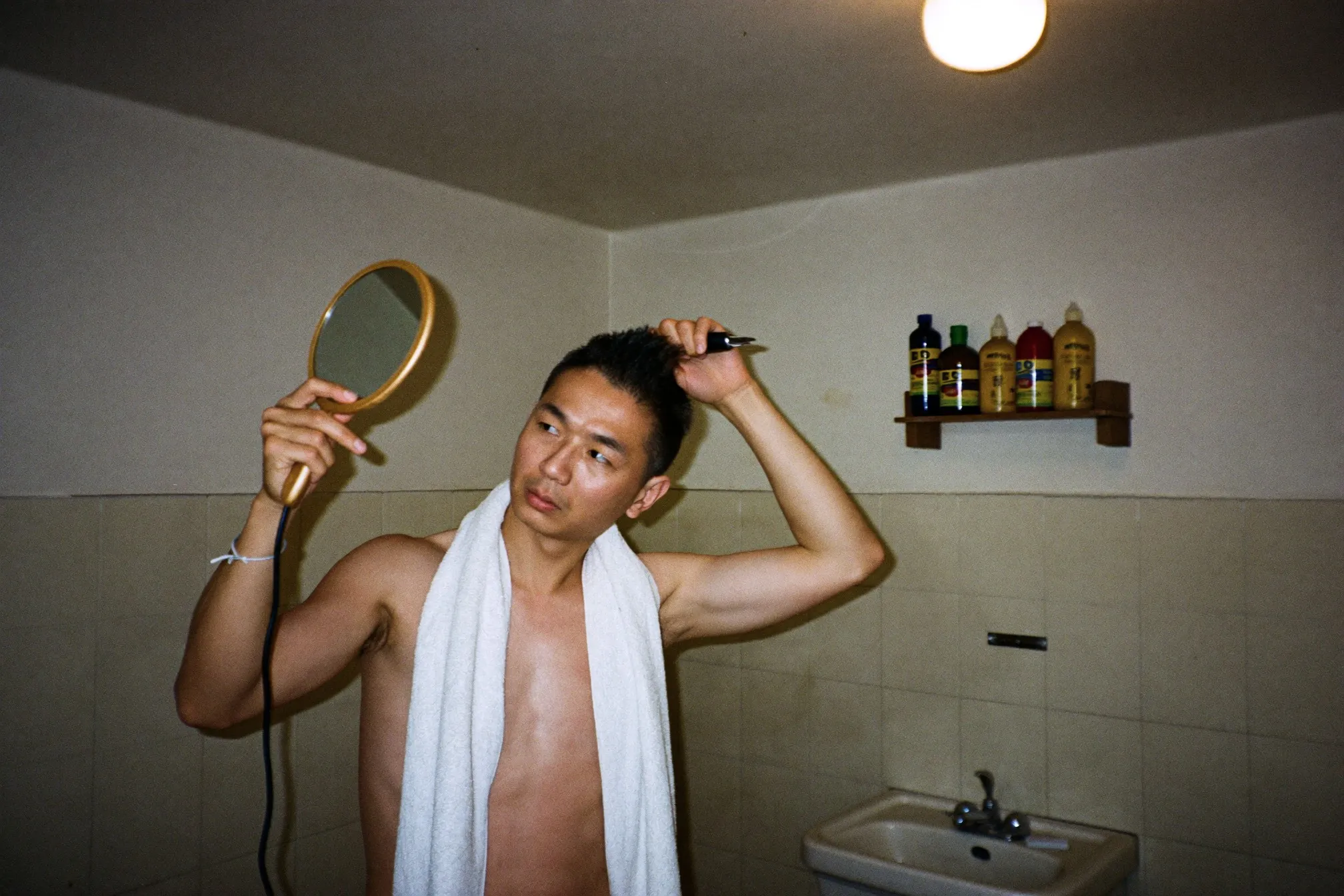 Man buzzing his own head with clippers over a bathroom sink in an HDB flat, using a handheld mirror