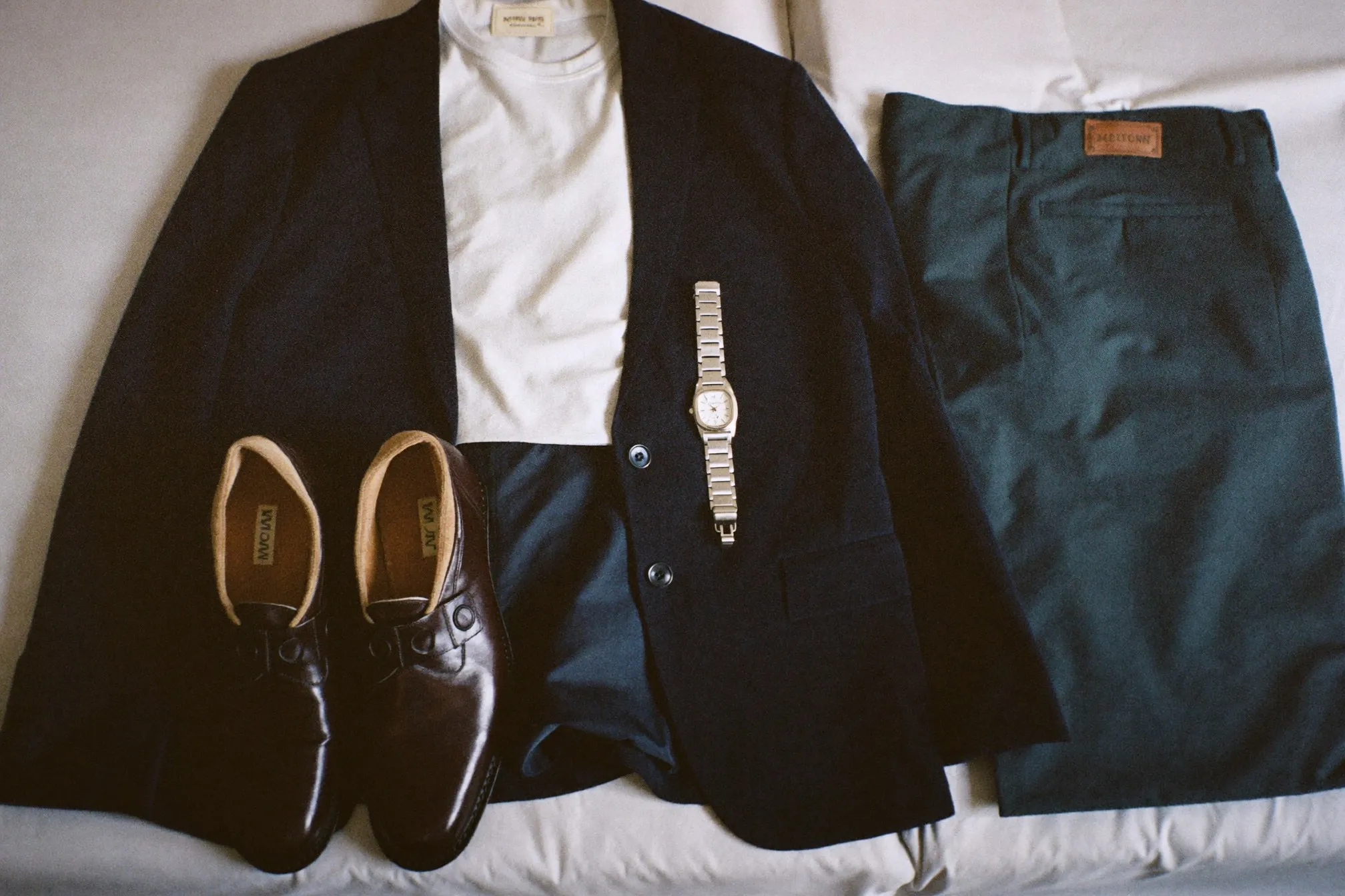 Flat lay of a navy blazer, white shirt, dark chinos, loafers and a steel watch on a bed