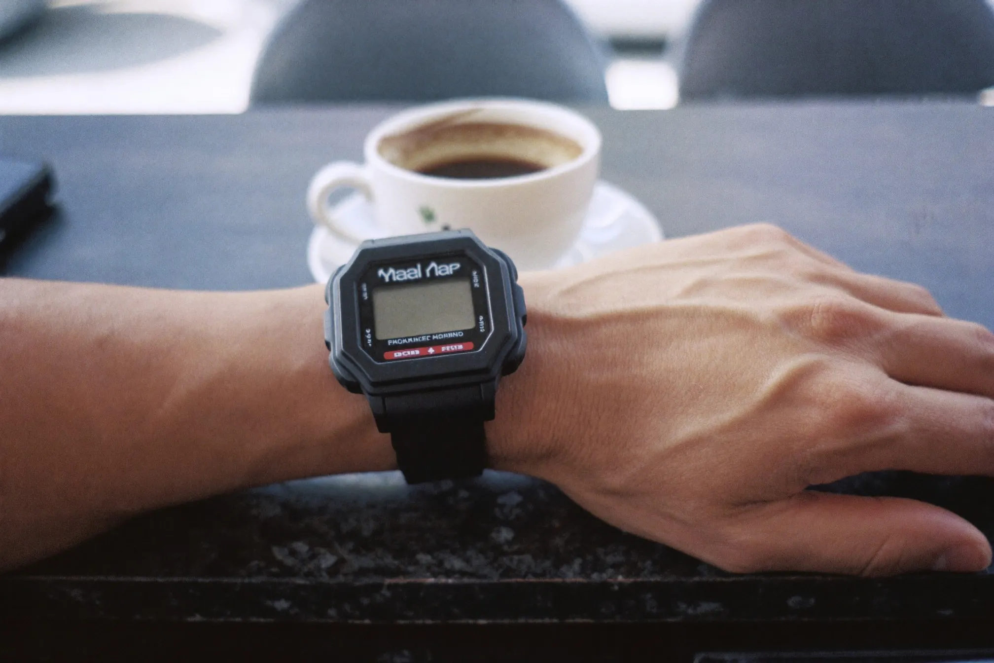 Black resin digital watch on a man's wrist resting beside a cup of kopi on a kopitiam marble table