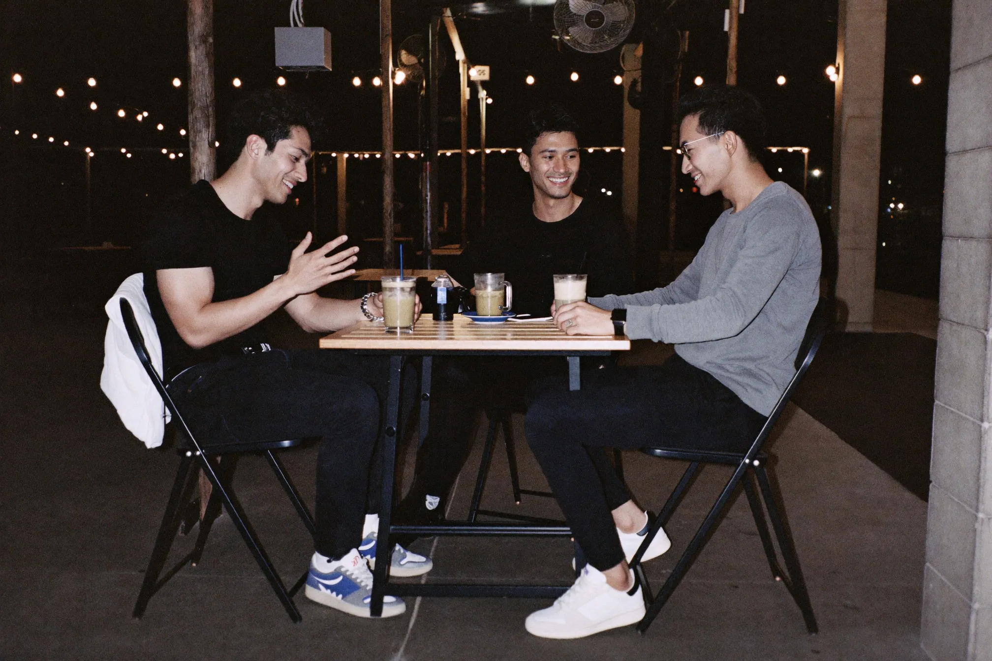 Two friends in fitted T-shirts, dark jeans and clean sneakers talking over teh tarik at an open-air mamak at night