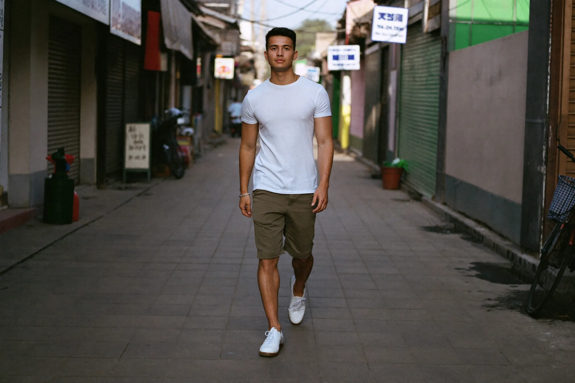 Man in a fitted white T-shirt, olive chino shorts and clean white sneakers walking along a covered shophouse walkway in Kuala Lumpur