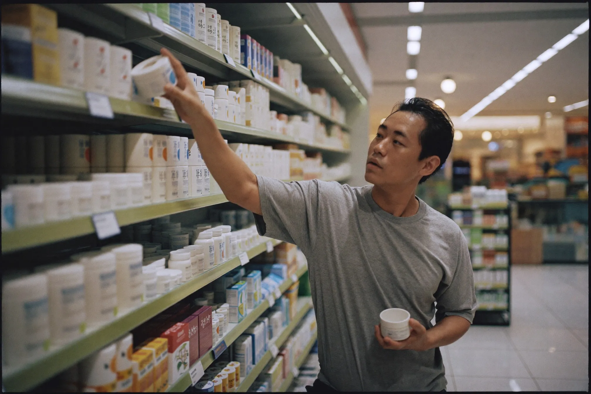 Man reaching for a plain white cream tub on a brightly lit pharmacy shelf in a shopping mall