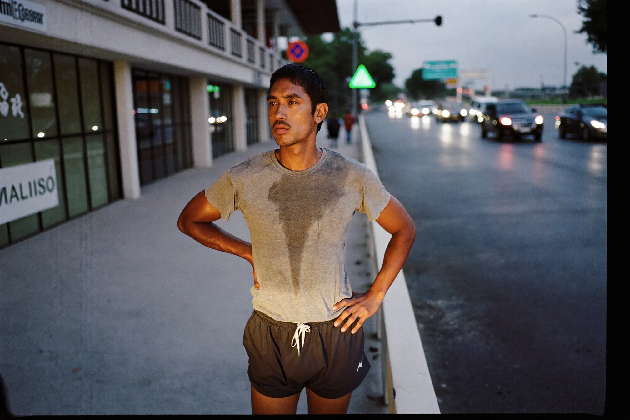 Man in running shorts and a damp T-shirt cooling down on a sheltered walkway after a humid evening run in Kuala Lumpur