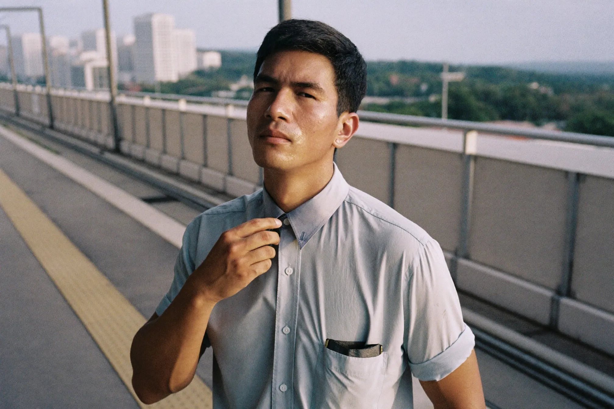 Man in a damp office shirt waiting on an open-air LRT platform in Kuala Lumpur during the afternoon heat
