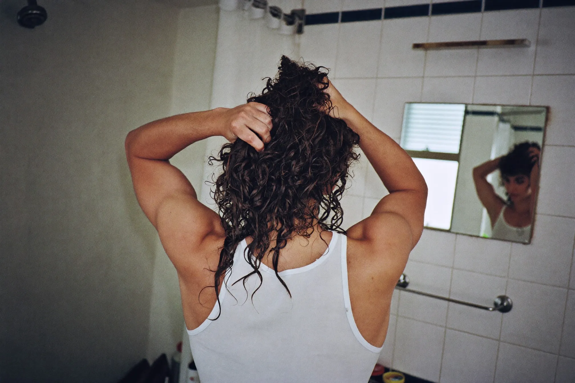 Young Malay man with loose natural curls scrunching product into wet hair in front of a bathroom mirror in a Kuala Lumpur flat