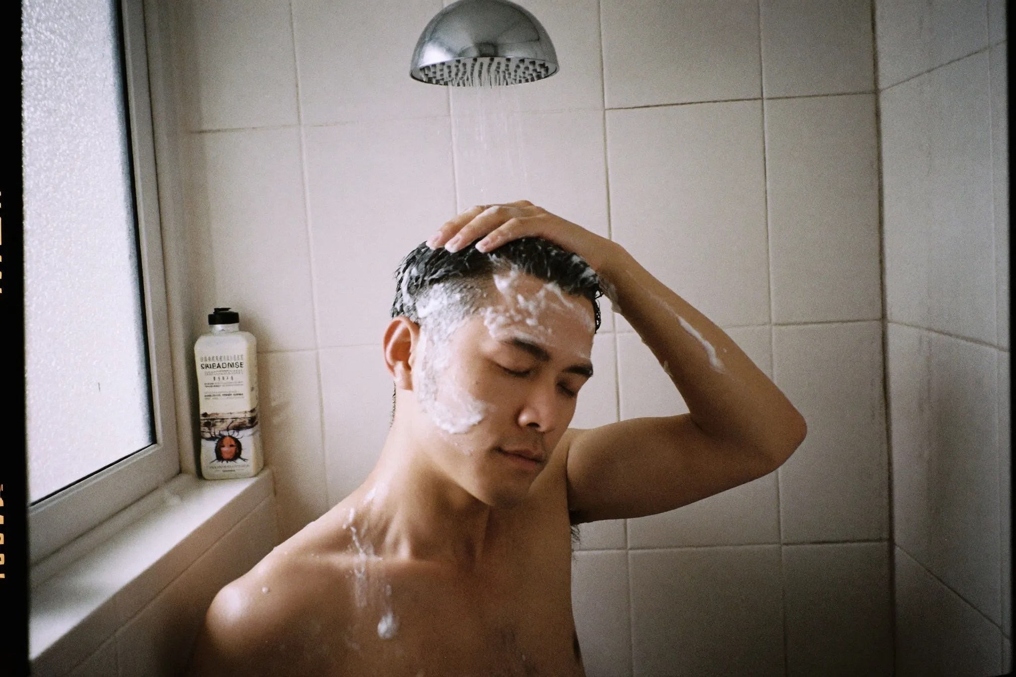 Man massaging shampoo into his scalp with his fingertips in a small tiled bathroom shower