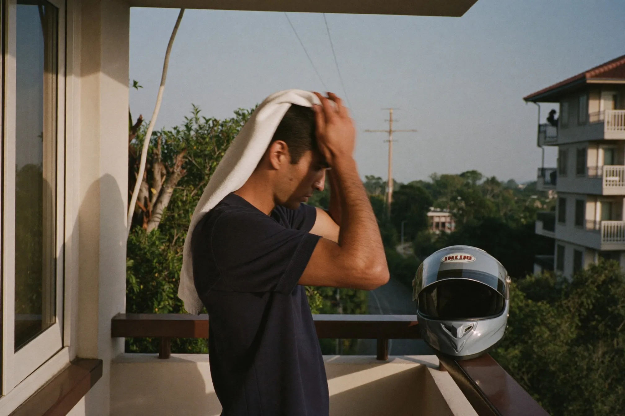 Man towel-drying his hair on an apartment balcony with an open-face helmet airing on the railing beside him