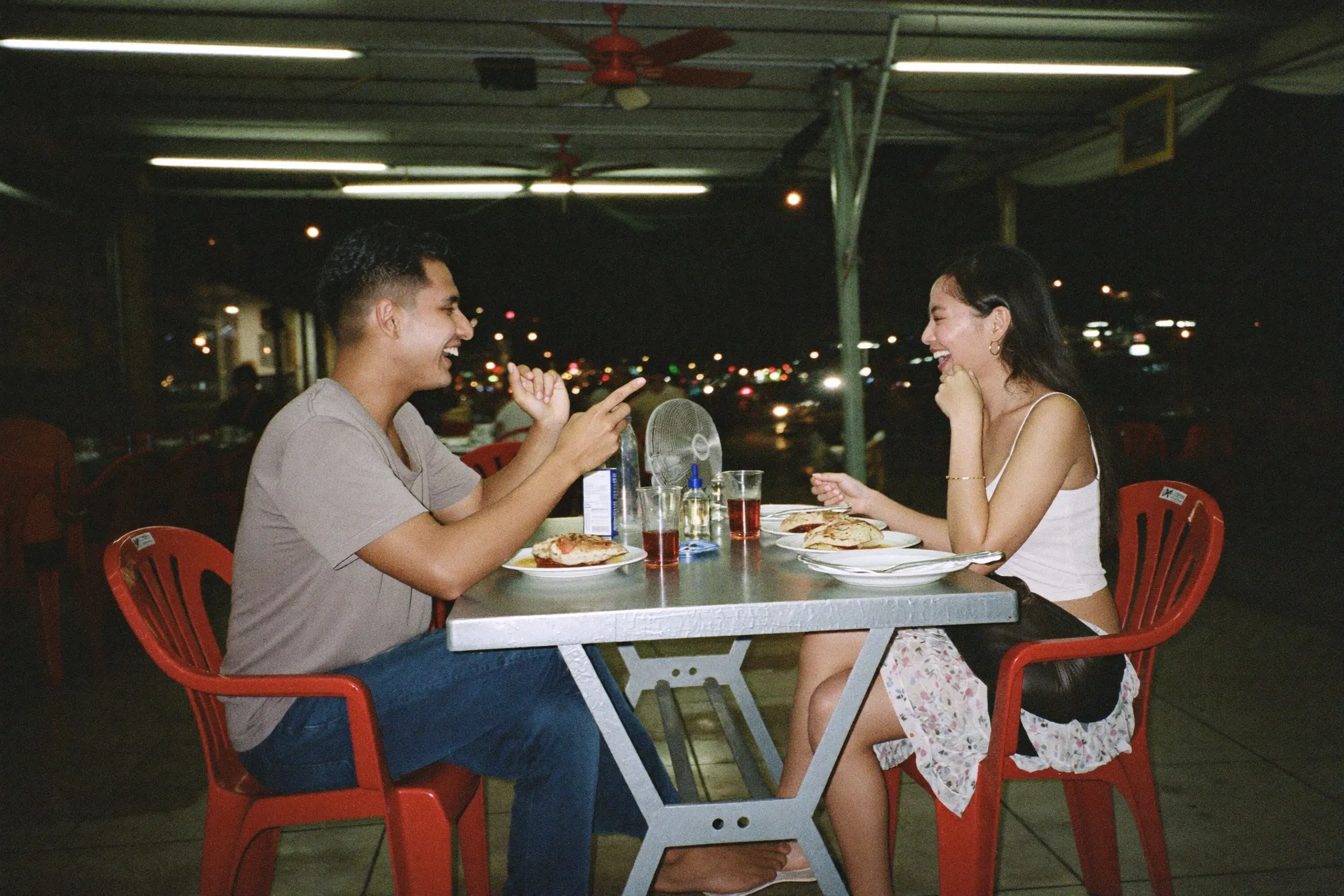 Two people sharing supper at a bright outdoor mamak table in Petaling Jaya at night