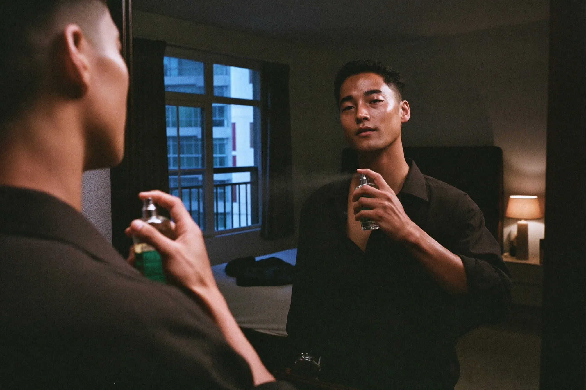 Man in a dark open-collar shirt spraying fragrance at his collarbone in a dim bedroom before going out