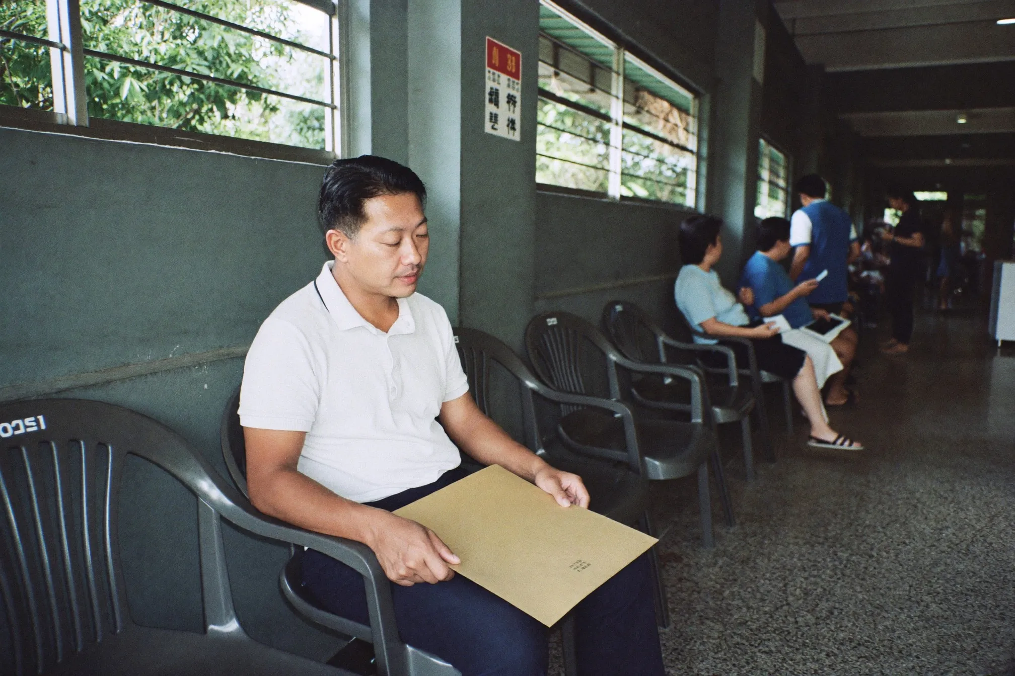 Man waiting on a row of chairs in a public health clinic corridor holding a folder of documents