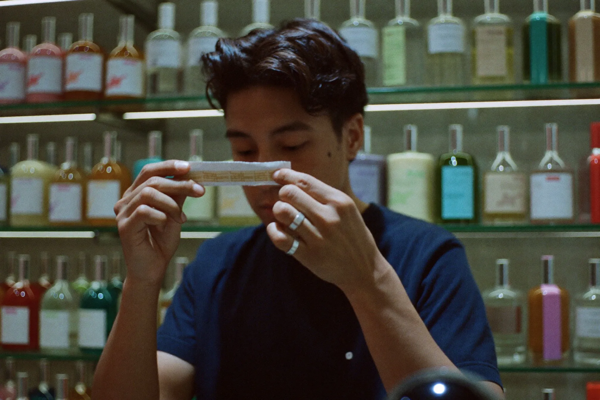 Man smelling a paper test strip at a department store fragrance counter, unbranded glass bottles on the shelves behind him