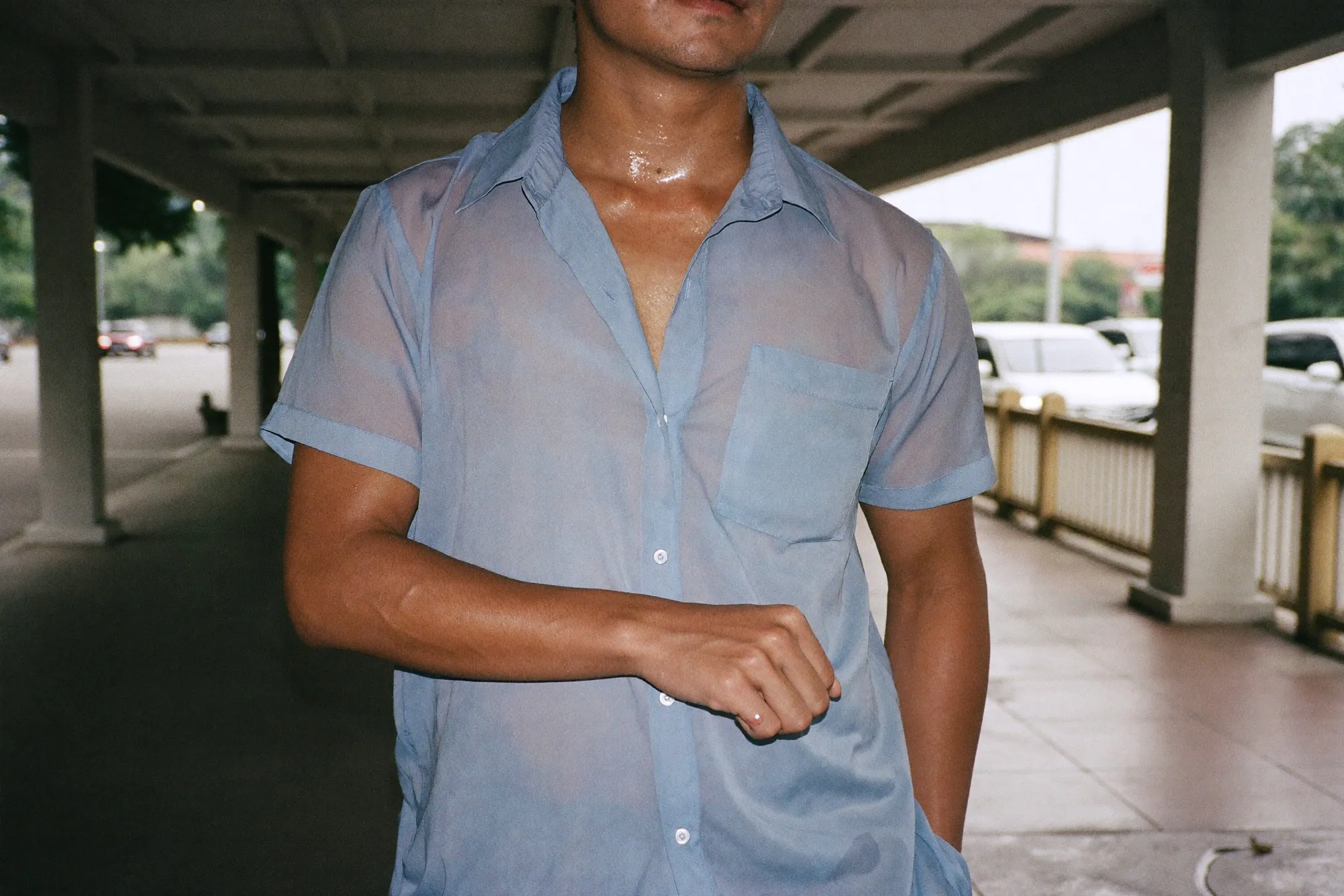 Man in a damp shirt standing under a covered walkway in heavy tropical humidity, scratching his forearm