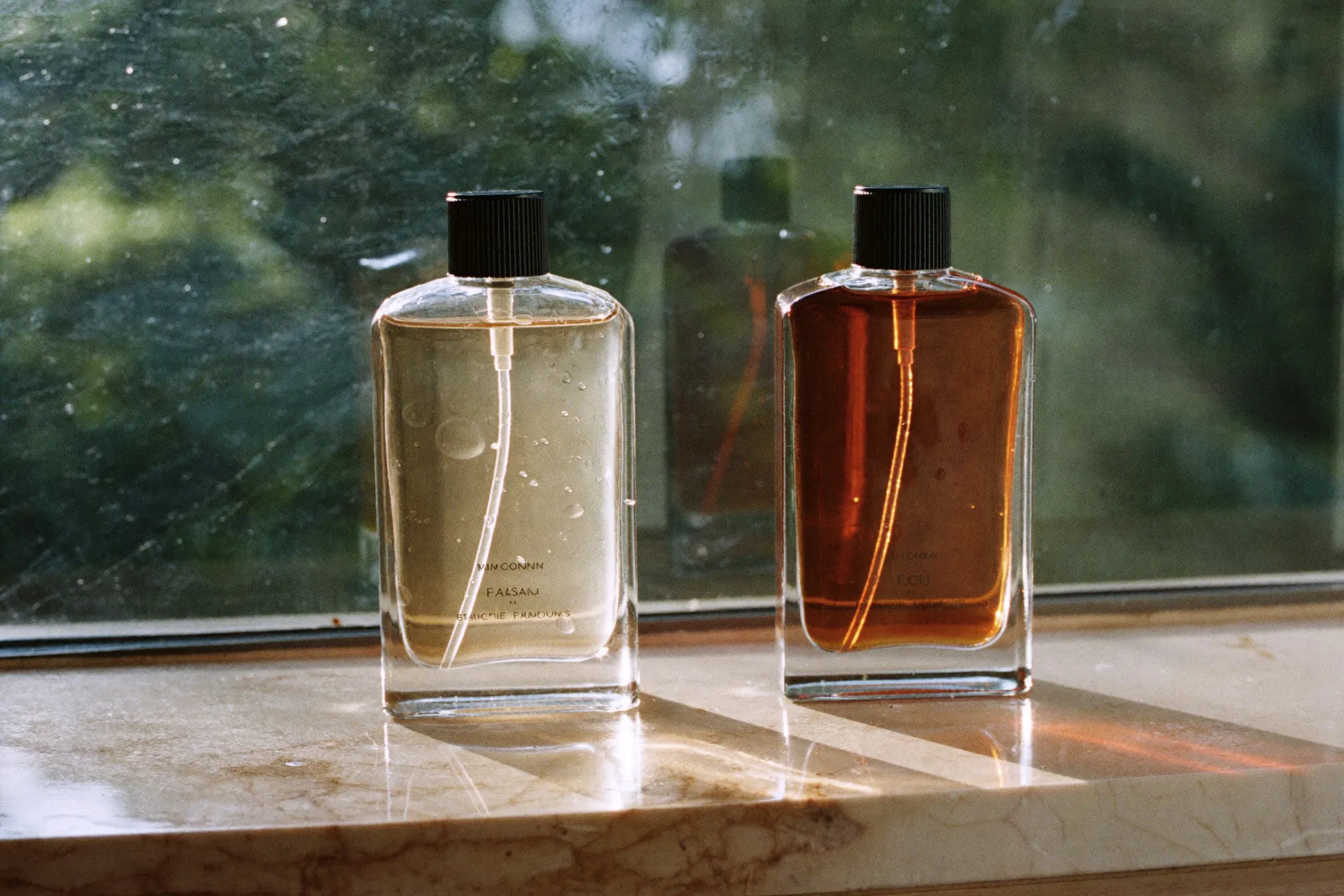 Two unbranded glass atomiser bottles side by side on a pale marble ledge, one clear and one deep amber