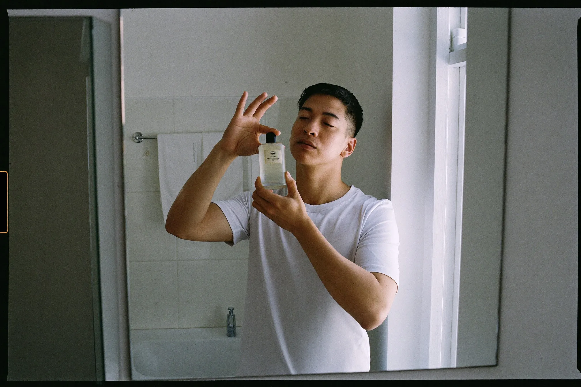 Man in a white t-shirt holding an unbranded frosted glass fragrance bottle up to the morning light at a bathroom mirror in a Singapore HDB flat
