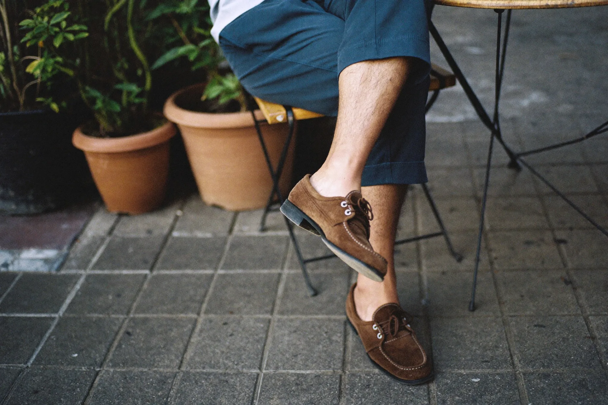 Man seated at an outdoor cafe table in Bangsar wearing cropped navy chinos and brown suede loafers with no-show socks
