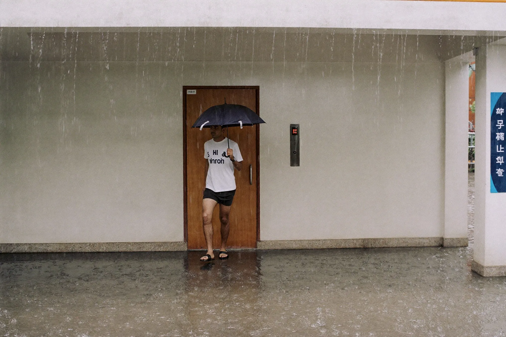 Man in shorts and black moulded rubber slides stepping onto wet pavement outside a lift lobby during a downpour