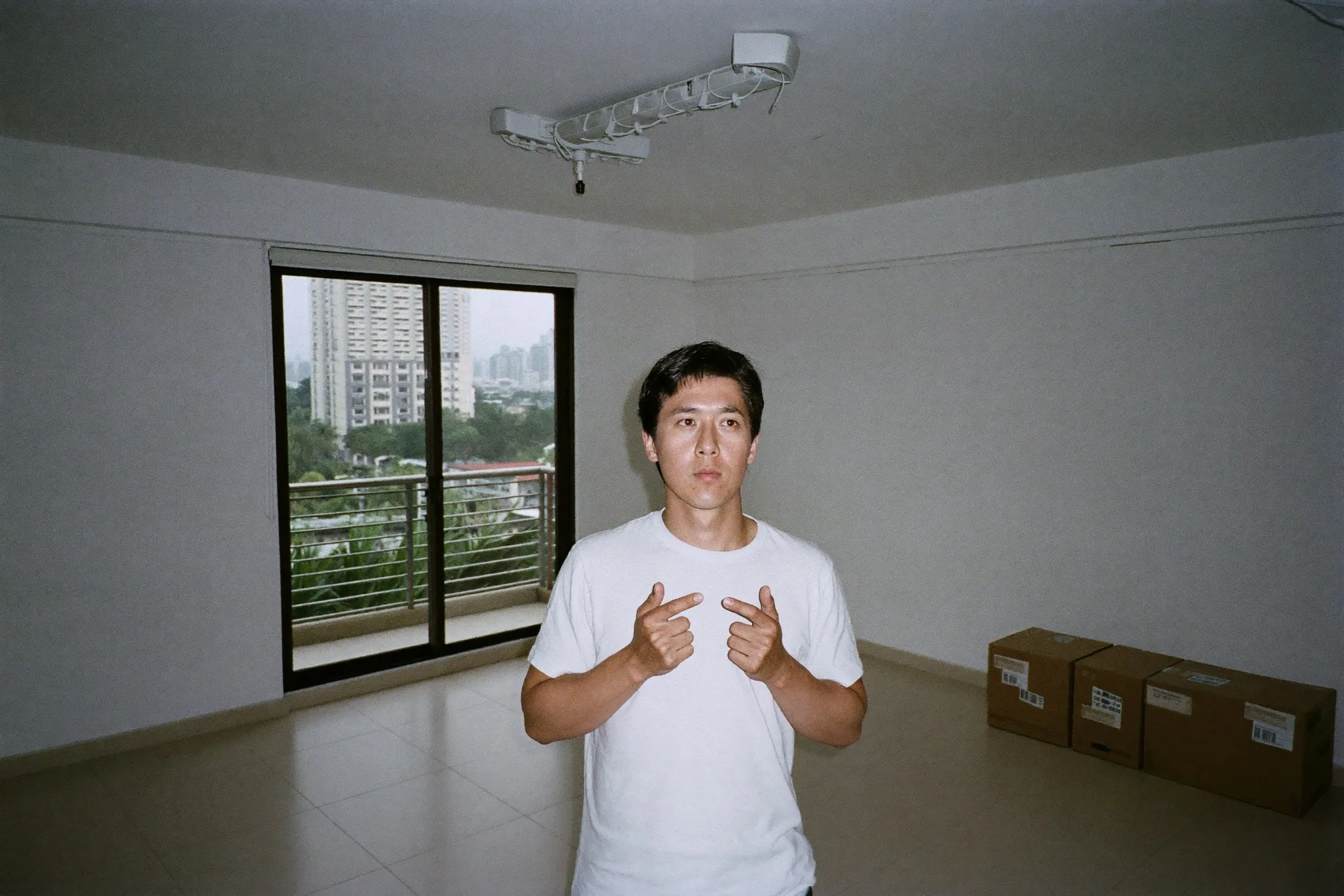 Young man standing in an empty unfurnished KL condo living room with moving boxes and the city skyline through the balcony doors
