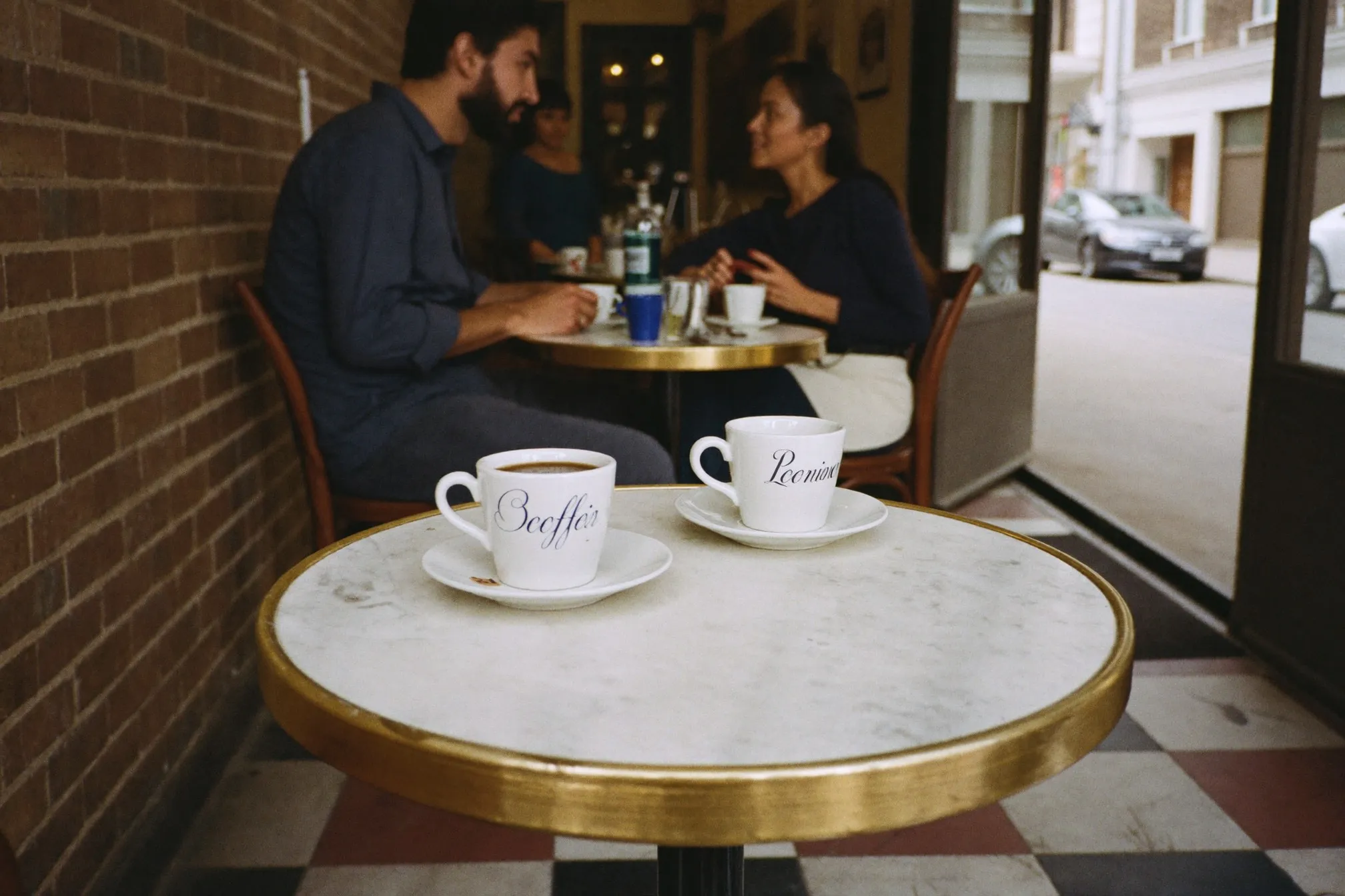 Two coffees on a small marble table at a narrow specialty cafe, two people talking in the background