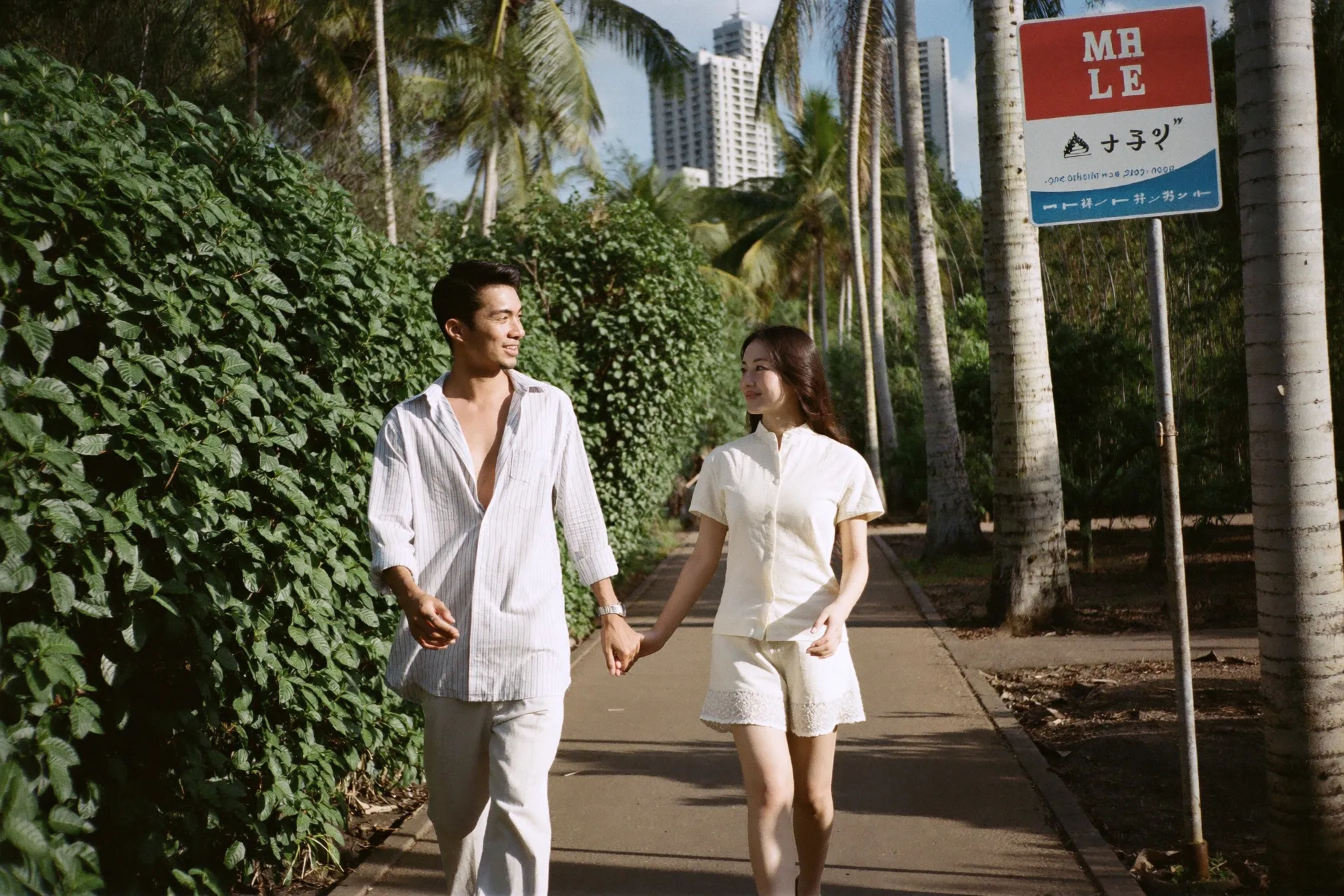 A man and a woman in their twenties walking and talking along a shaded park path in Kuala Lumpur on a bright afternoon