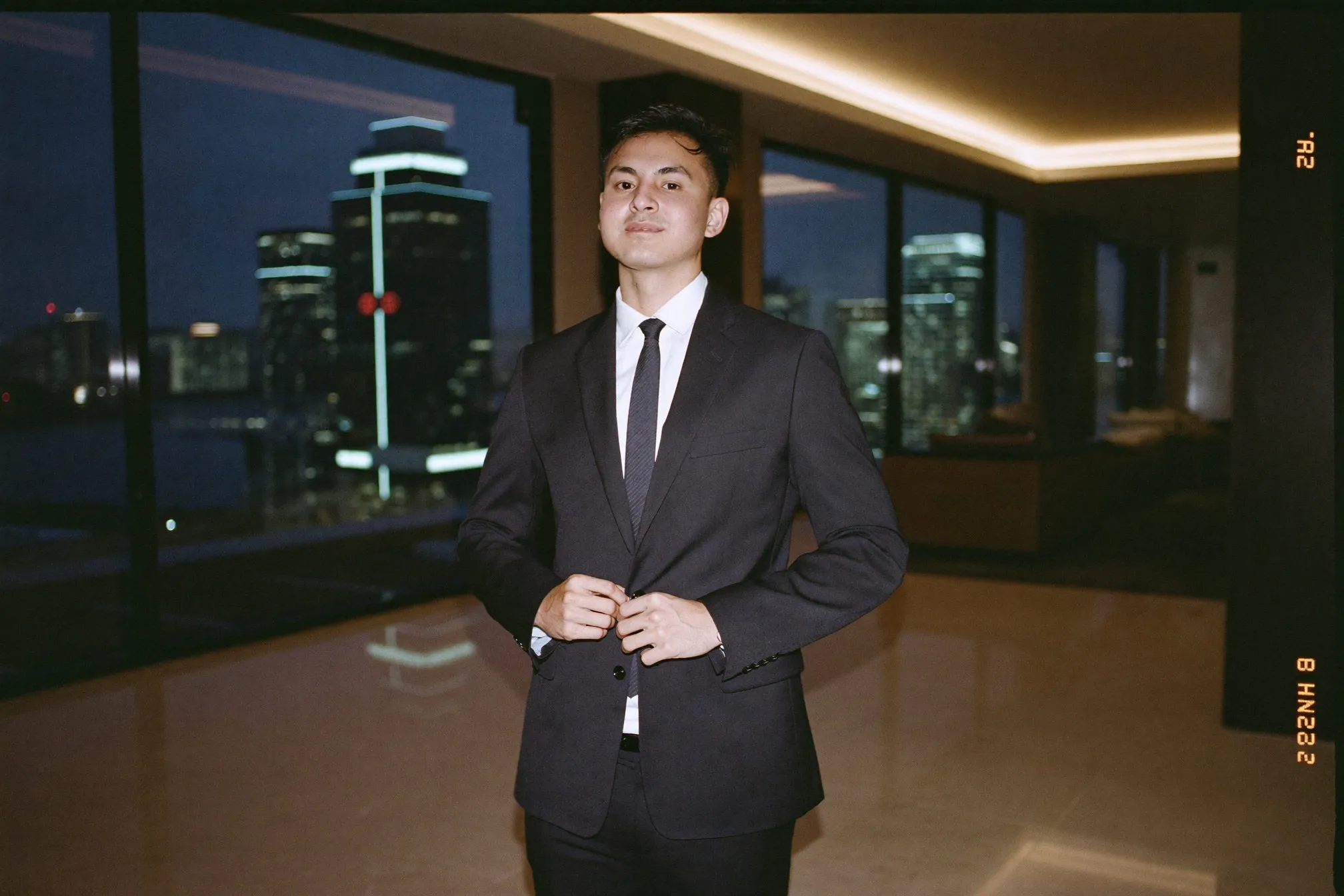 Man in a dark navy suit and tie fastening his jacket button in a hotel lobby in Kuala Lumpur at dusk