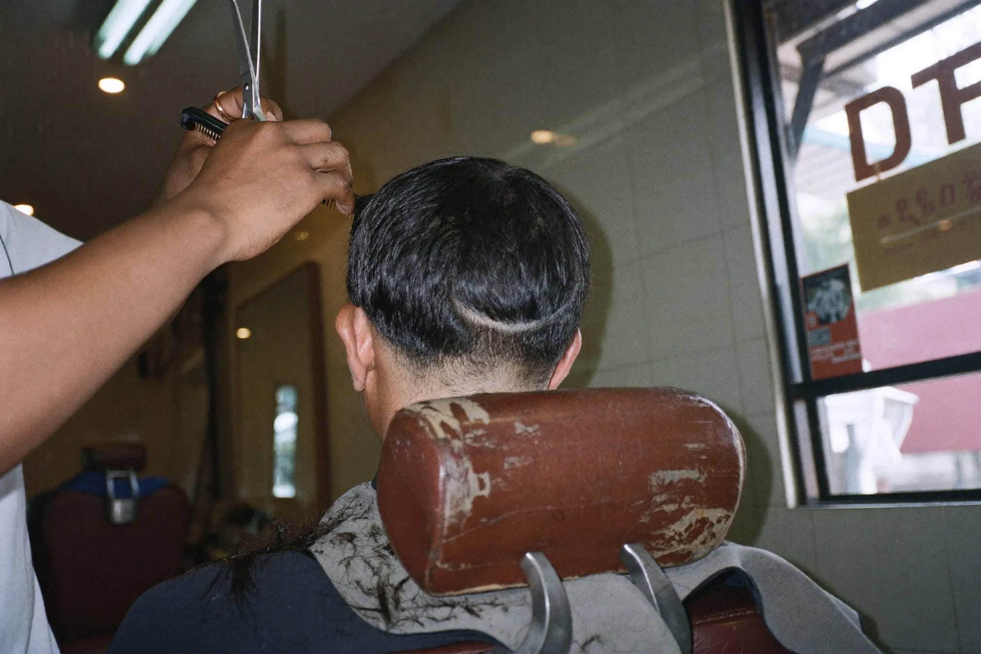 Barber point cutting the textured top of a French crop with scissors on a Malay man seated in a barber chair